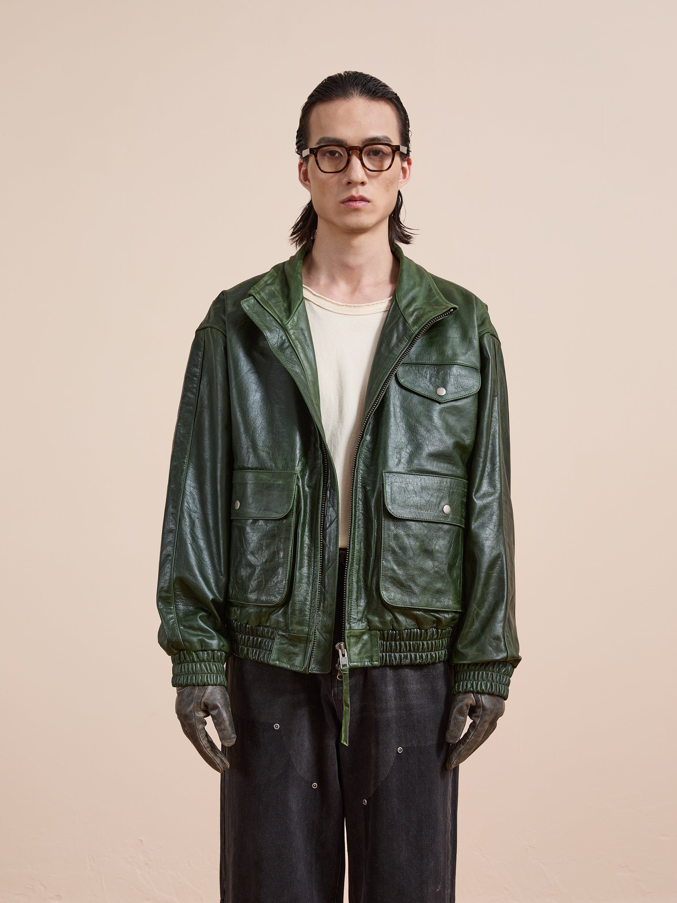 A person with long hair, glasses, a white shirt, FOUND’s Forest Leather Bomber Jacket in green, black pants, and gloves stands facing forward against a plain beige background.
