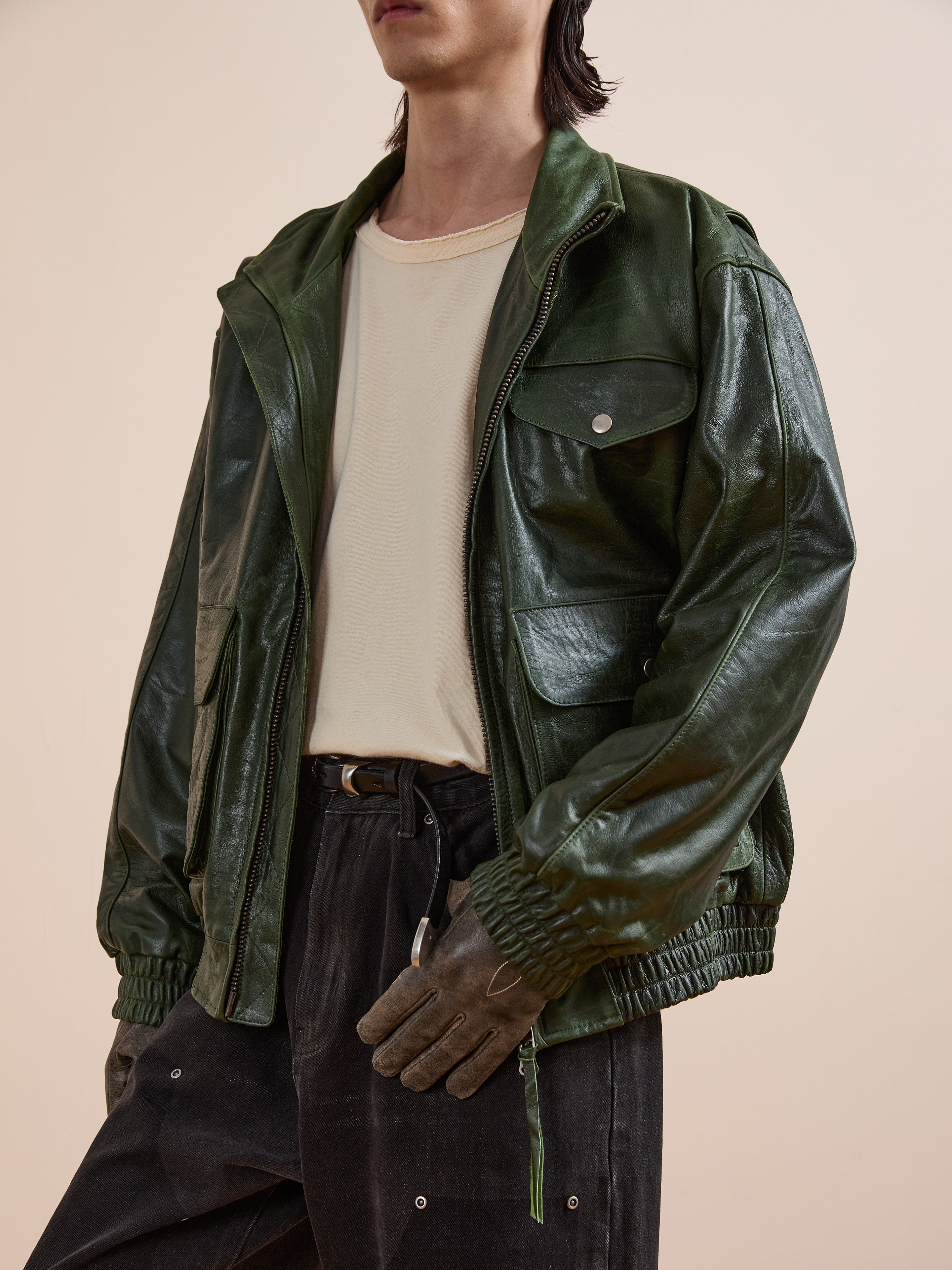 A person wears the FOUND Forest Leather Bomber Jacket in shiny green over a beige t-shirt, paired with dark pants and brown gloves, posing against a plain light background.
