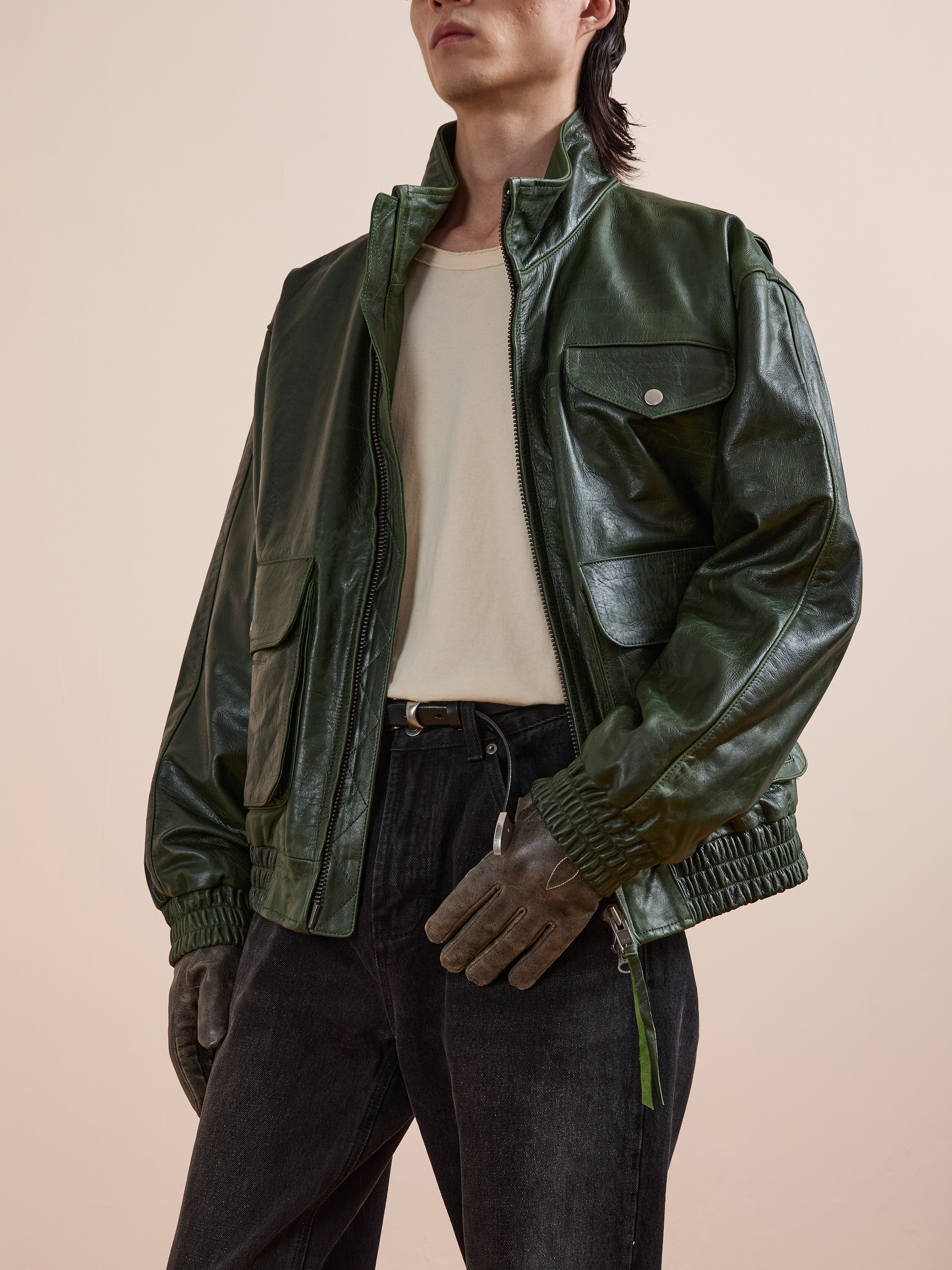A person wearing the FOUND Forest Leather Bomber Jacket, a white t-shirt, dark jeans, and gray gloves stands against a plain light background.