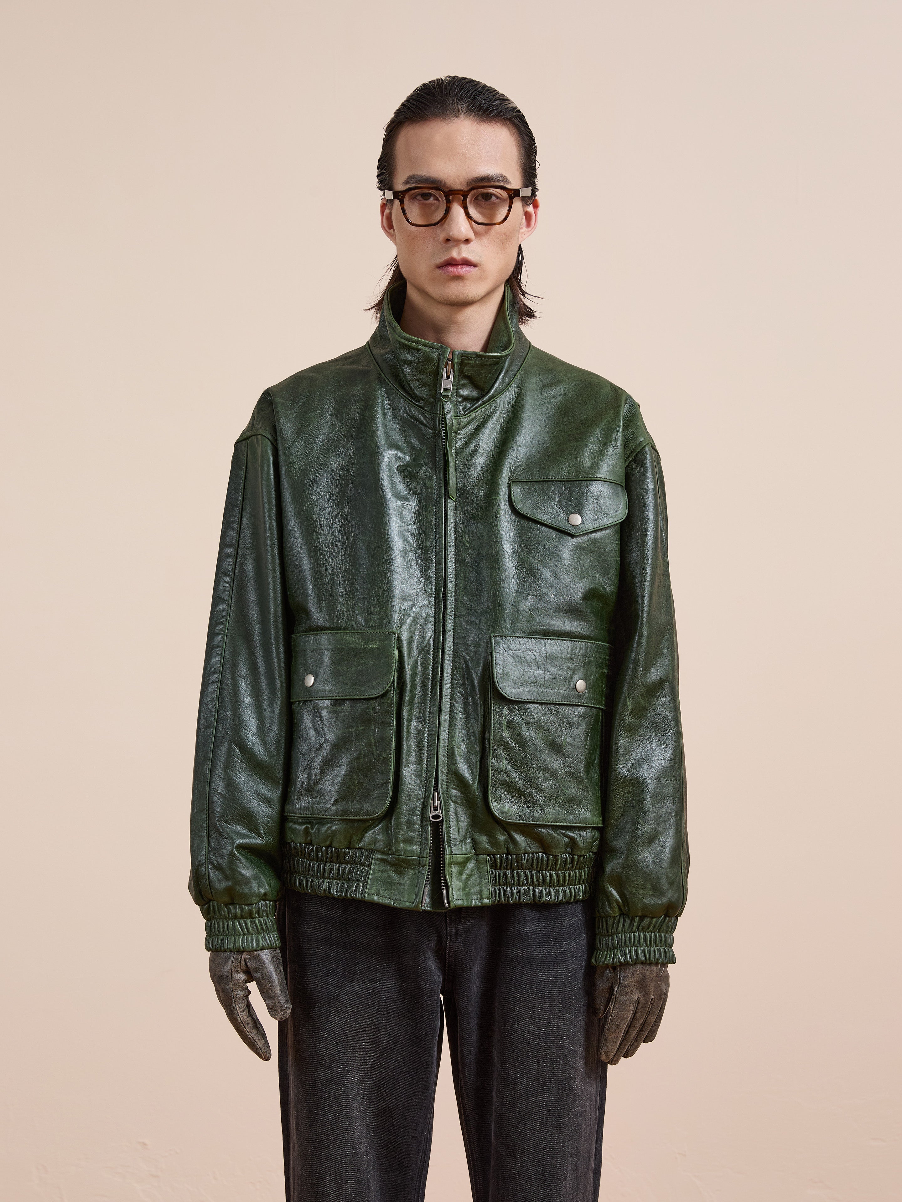 A person wearing glasses, the FOUND Forest Leather Bomber Jacket with front pockets, dark pants, and brown gloves stands against a plain beige background.