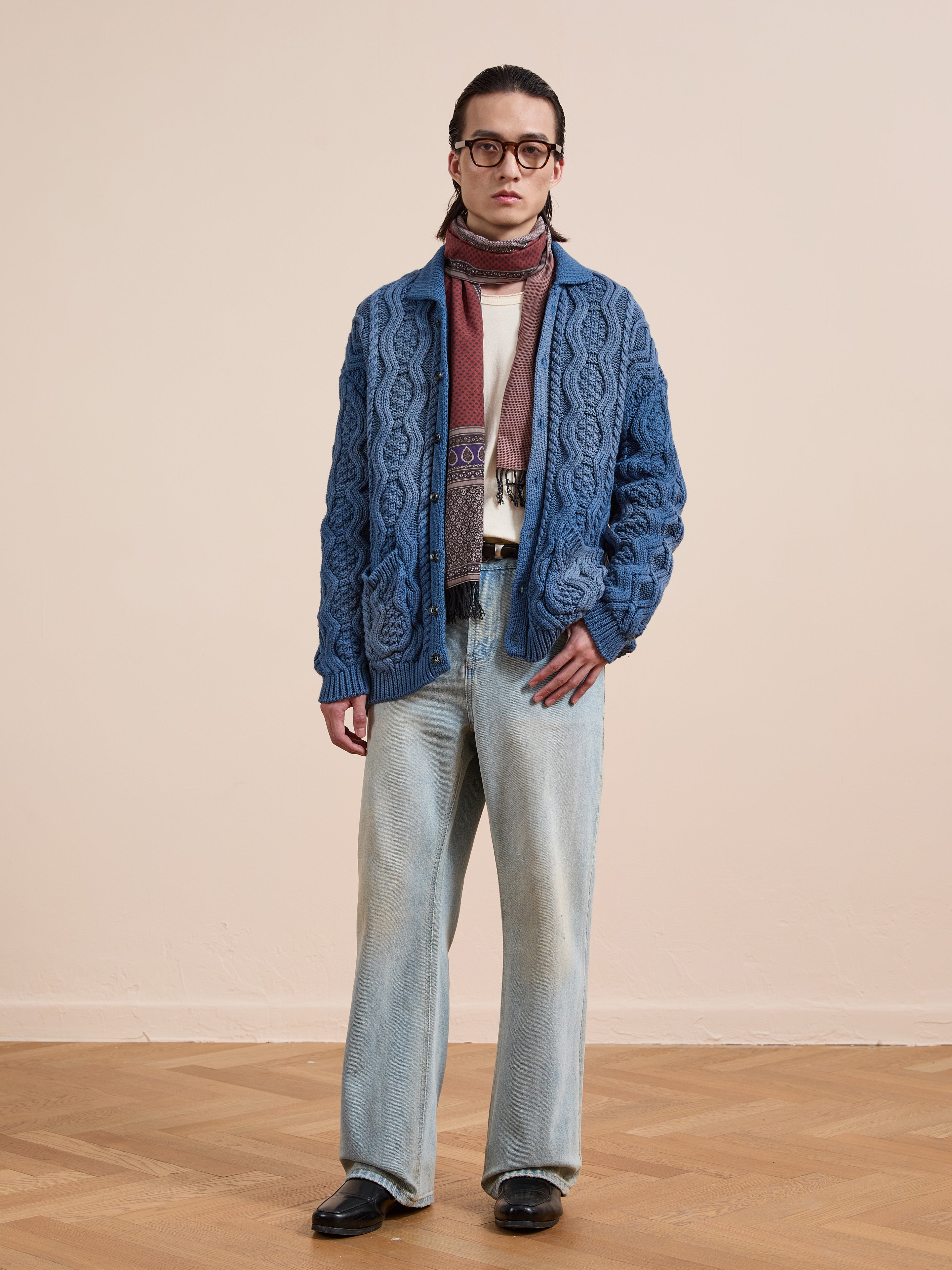 A person in glasses wears the Found Cable Knit Buttoned Cardigan (navy, unisex) with a patterned scarf, white shirt, light blue jeans, and black shoes, standing on a wooden floor against a plain beige wall.