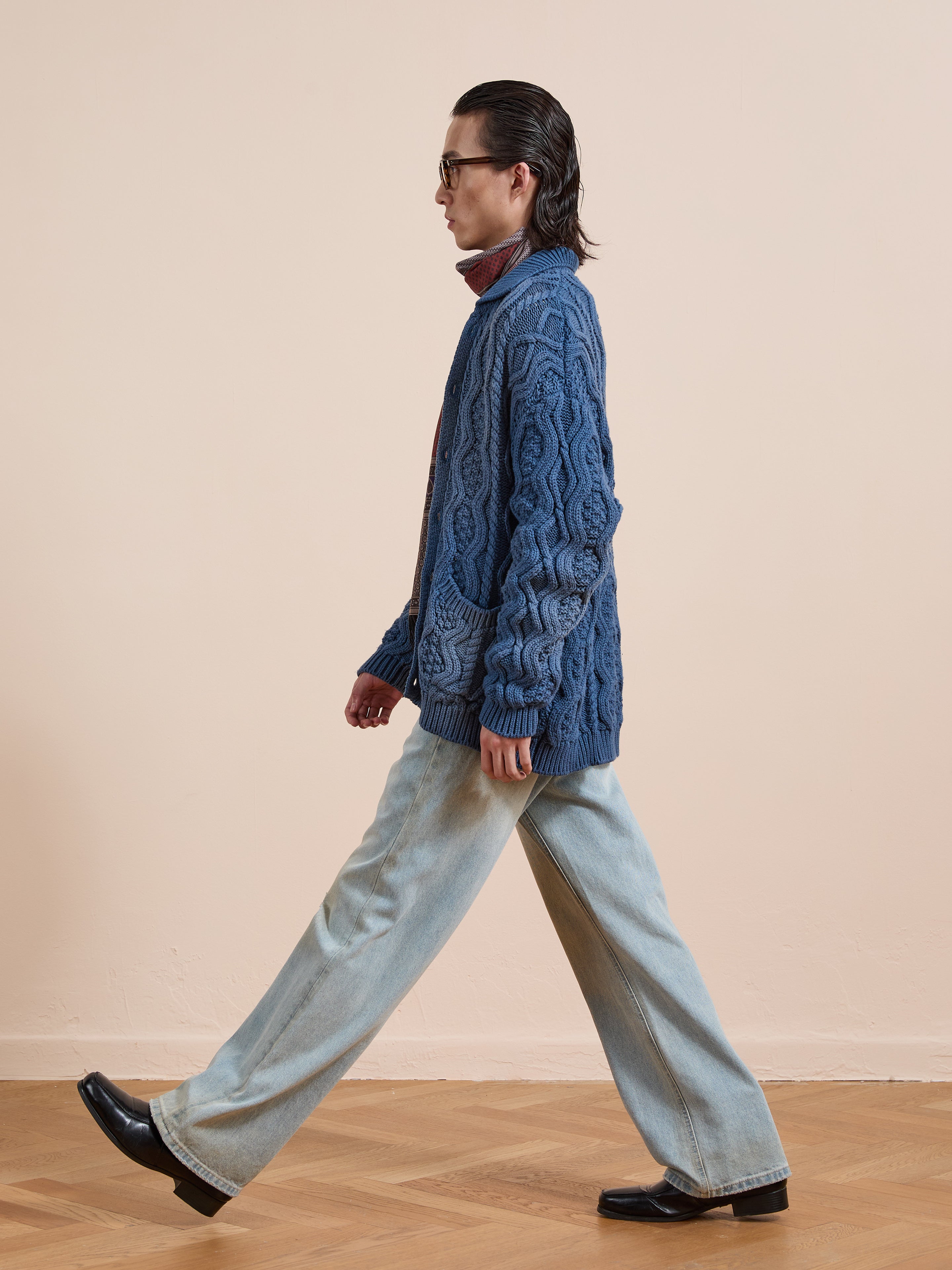 A person with long hair and glasses walks indoors in a navy Cable Knit Buttoned Cardigan by Found, light blue wide-leg jeans, and black shoes against a beige background.