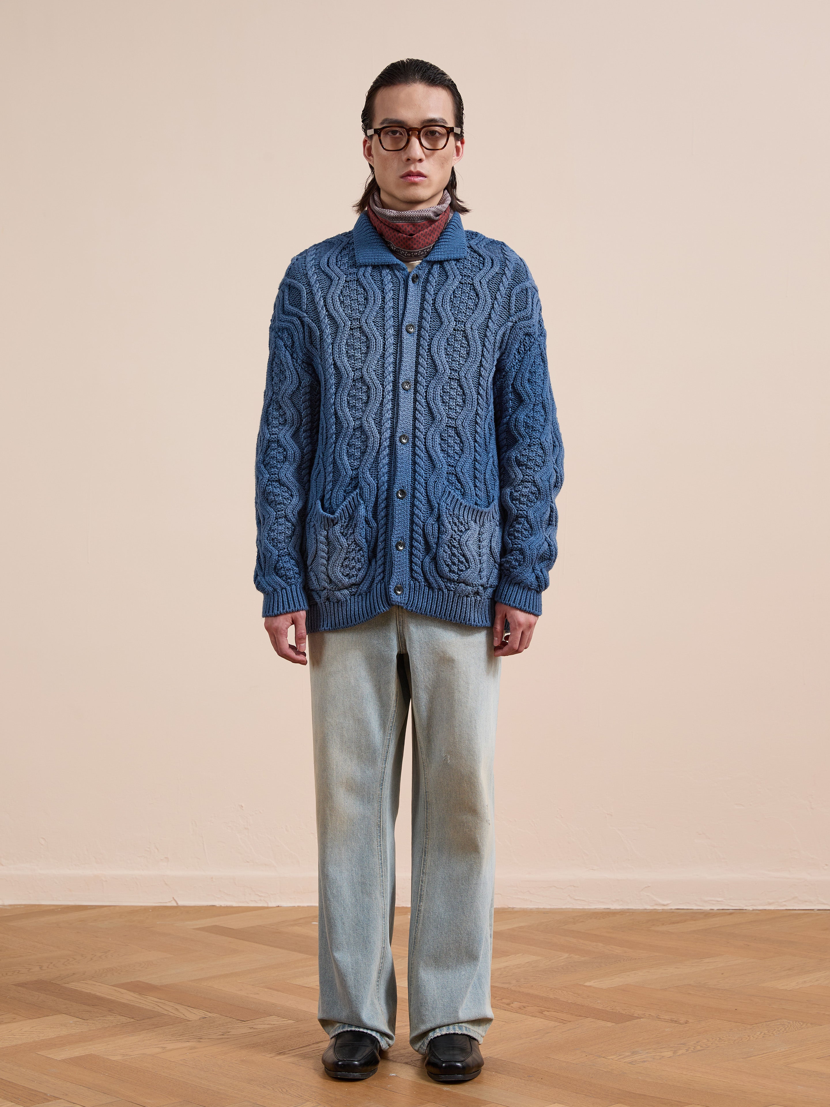 Indoors on a wooden floor, a person wearing glasses sports the Found Cable Knit Buttoned Cardigan, light-wash jeans, black shoes, and a patterned scarf.