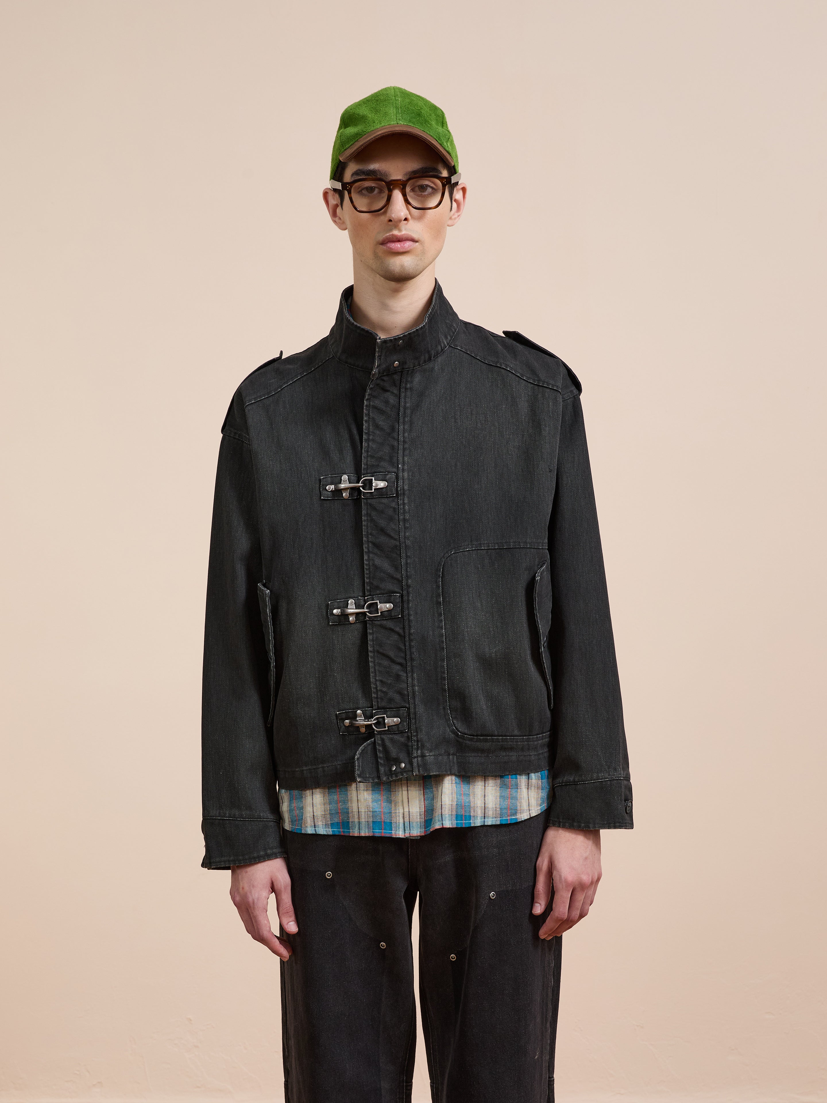 A person in a green cap, glasses, the Found Utility Clasp Work Jacket over a plaid shirt, and black pants stands against a plain beige background.