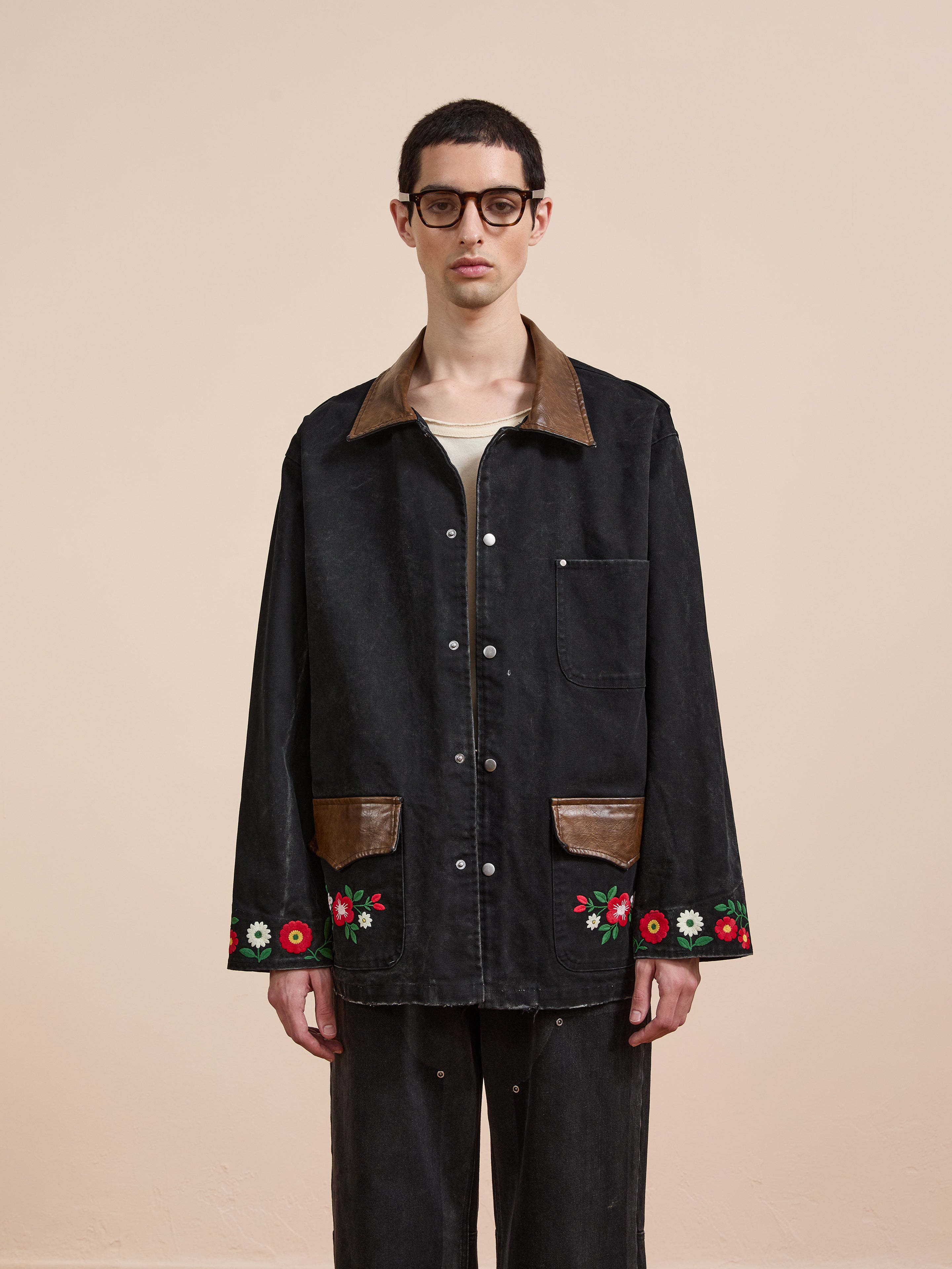 A person wearing Found's Canvas Embroidered Work Jacket, featuring a black canvas body, brown collar and pocket flaps, and red floral embroidery, stands against a plain beige background.