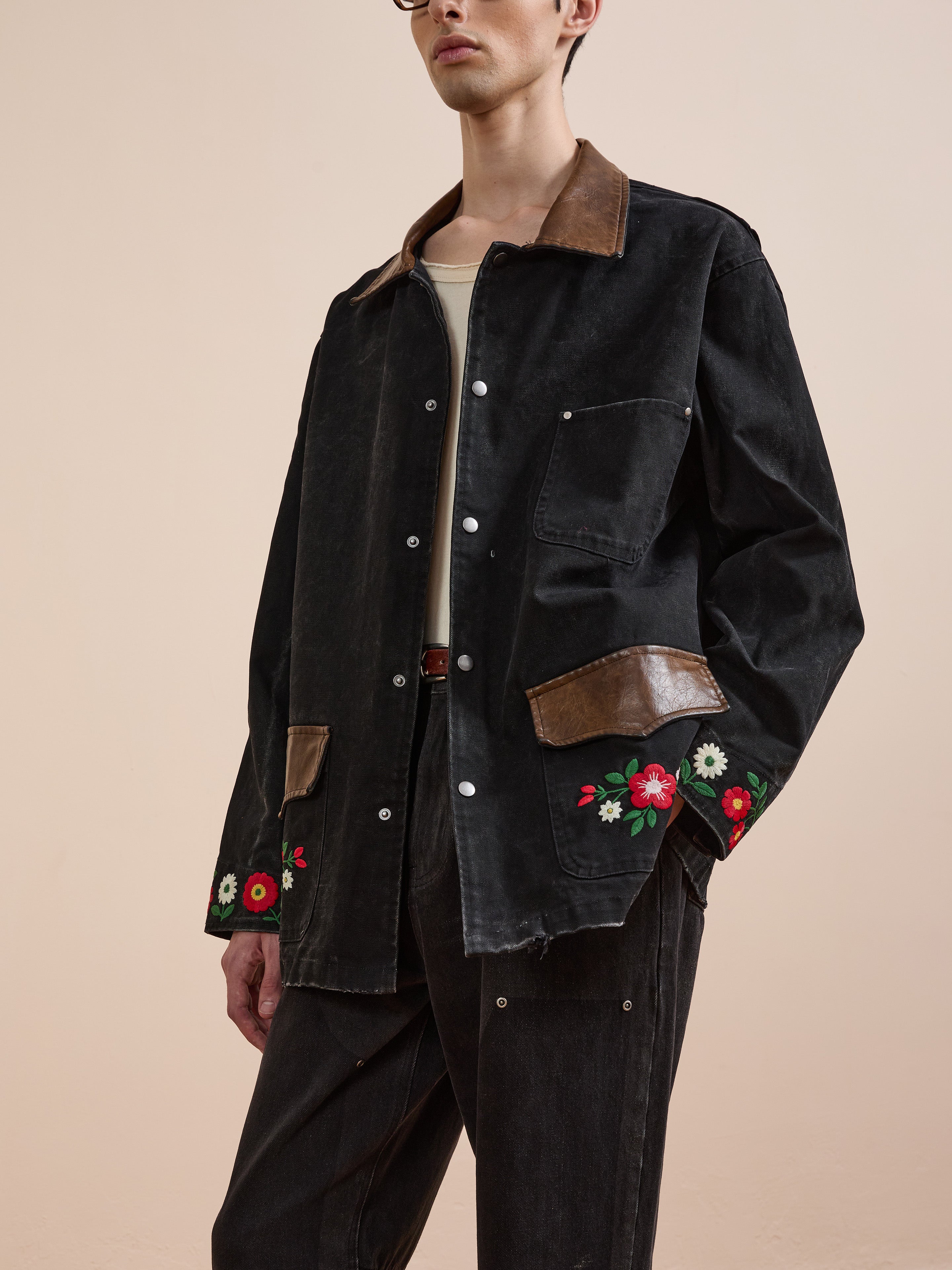 A person wears the Found Canvas Embroidered Work Jacket—black canvas with brown leather accents and floral embroidery—paired with black pants and a cream shirt for a stylish, unisex relaxed fit.