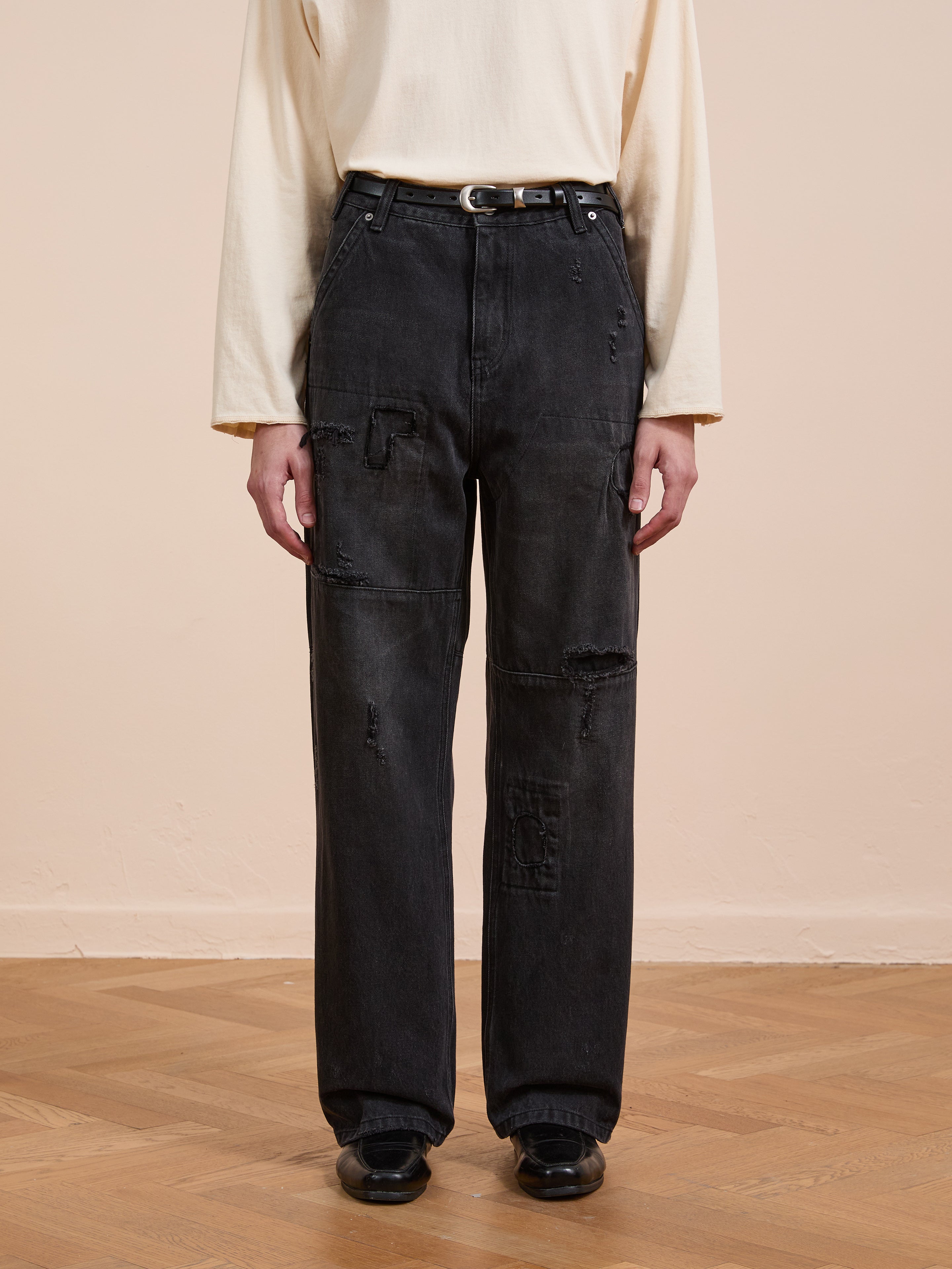 A person stands on a wooden floor against a plain background, wearing FOUND's Painter Repaired Jeans with patches, a long-sleeve beige shirt, black belt, and black shoes.