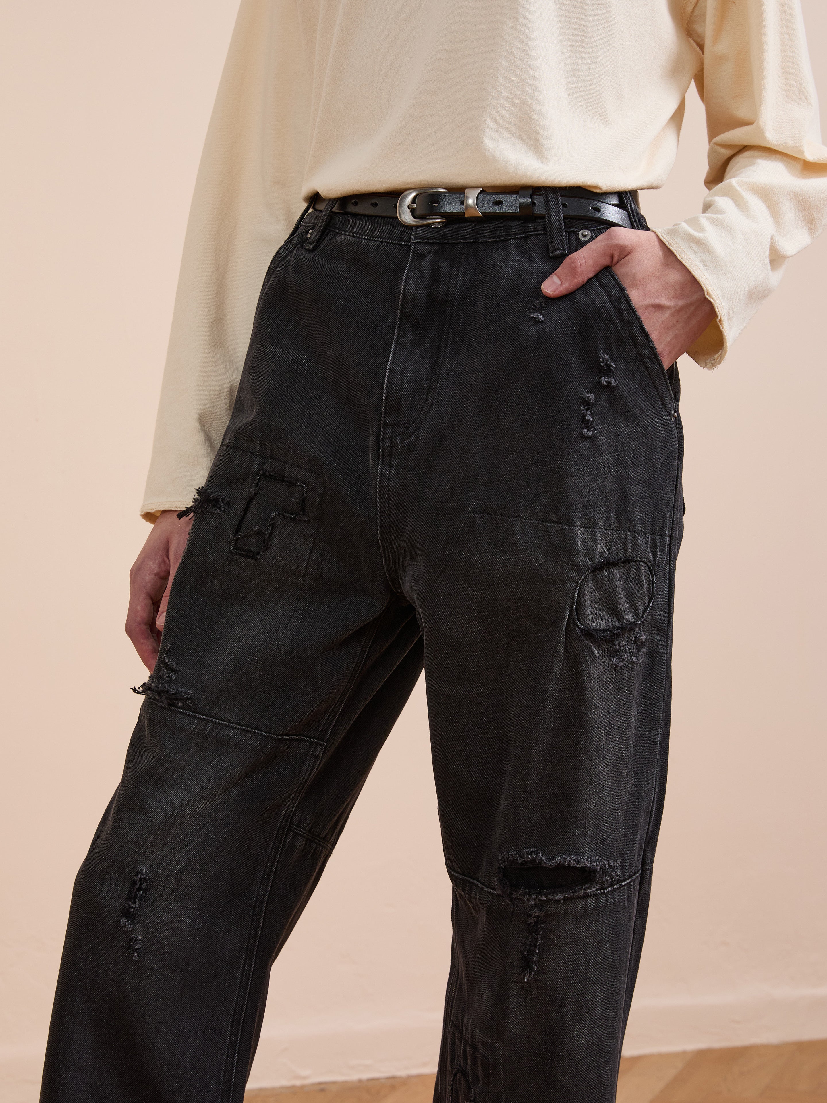 A person wears a long-sleeve cream shirt tucked into FOUND Painter Repaired Jeans, high-waisted black patchwork denim styled with a black belt. One hand is in the jeans pocket.