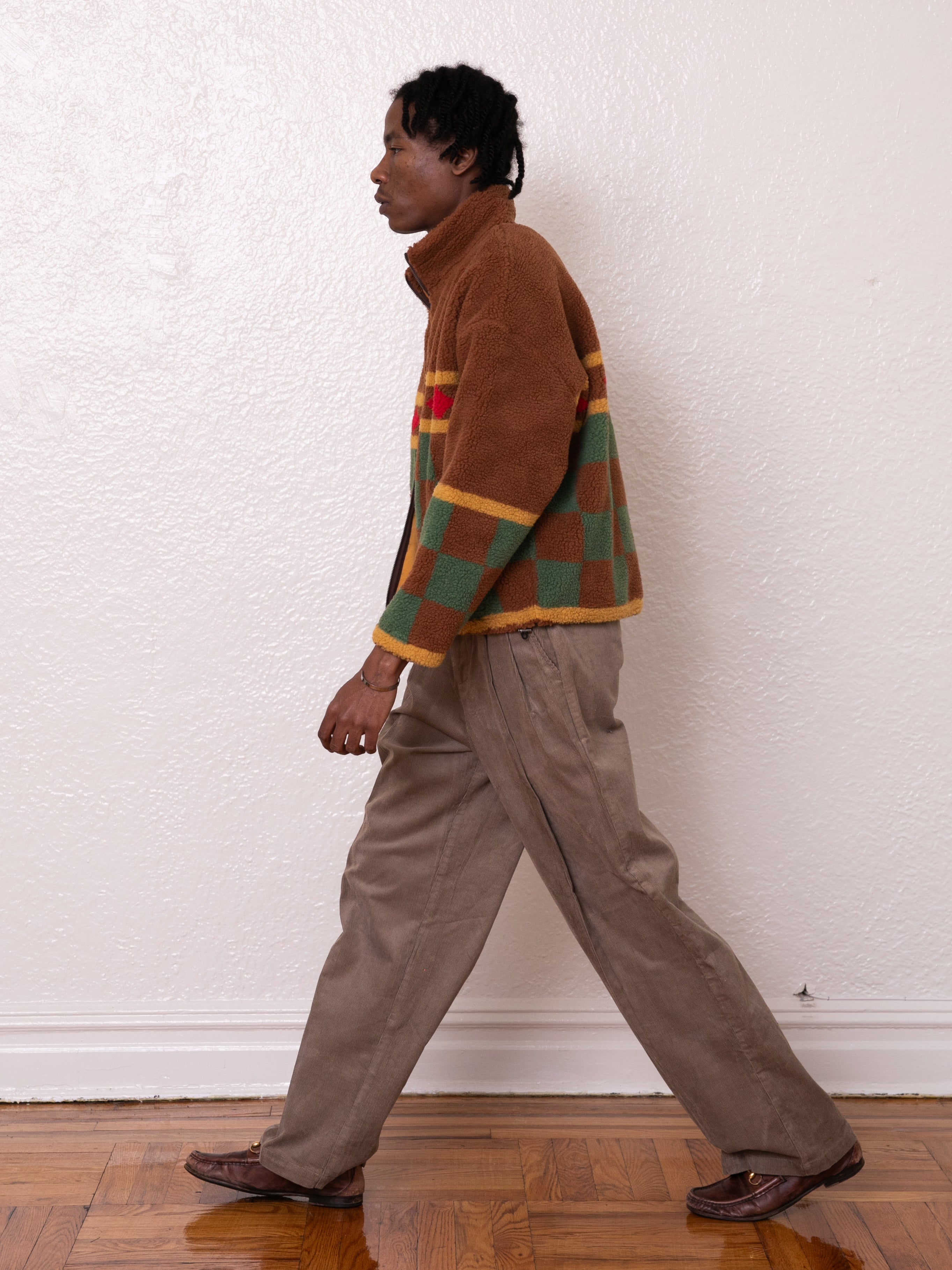 A person strolls indoors in the Checkered Zip-Up Sherpa by FOUND, along with brown pants and loafers, moving across a wooden floor against a plain wall.