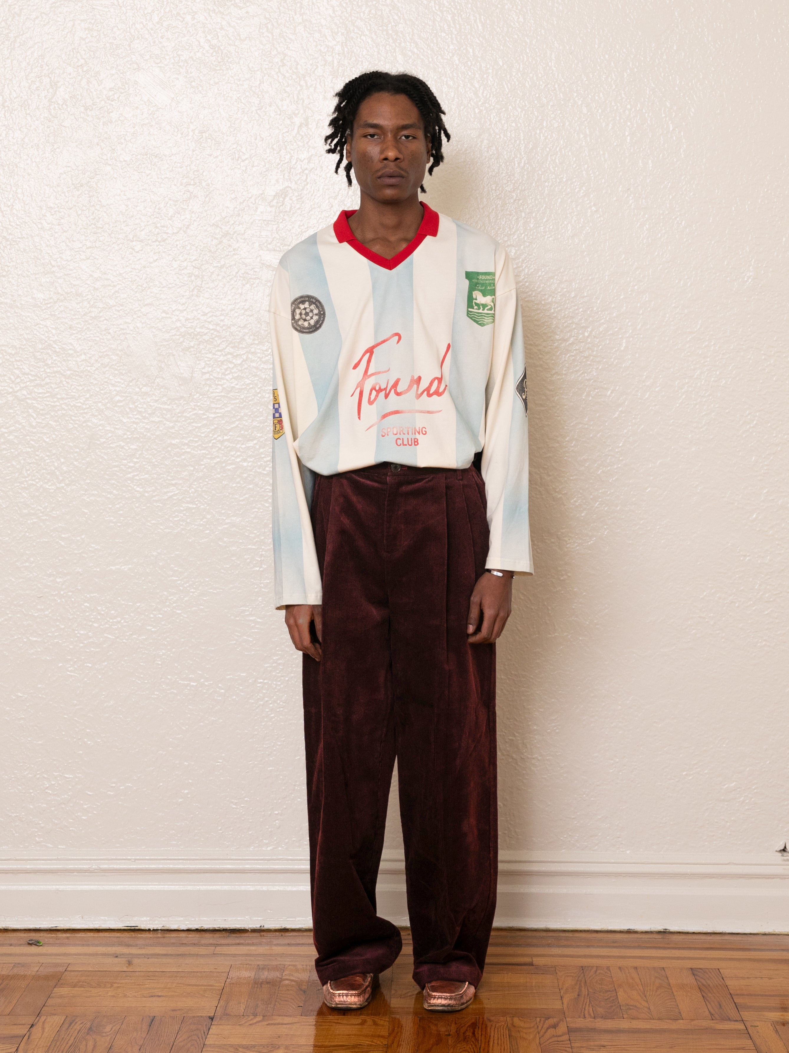 A person stands against a white wall wearing a long-sleeve shirt with "Found" text, Corduroy Pleated Trousers by FOUND, and sandals. The floor is wooden.