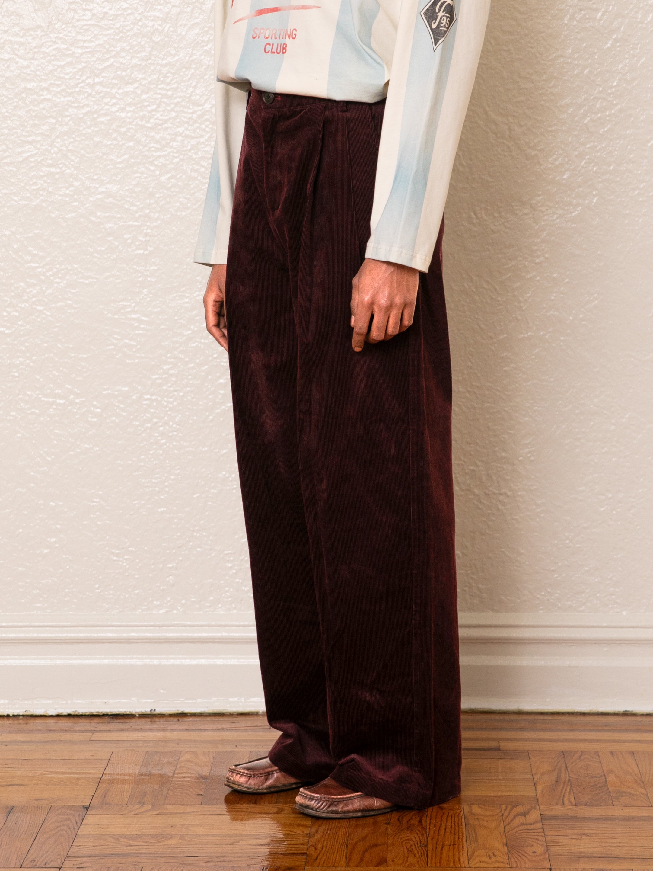 A person is on a wooden floor, wearing a light shirt and high-rise wide-leg Corduroy Pleated Trouser by FOUND.