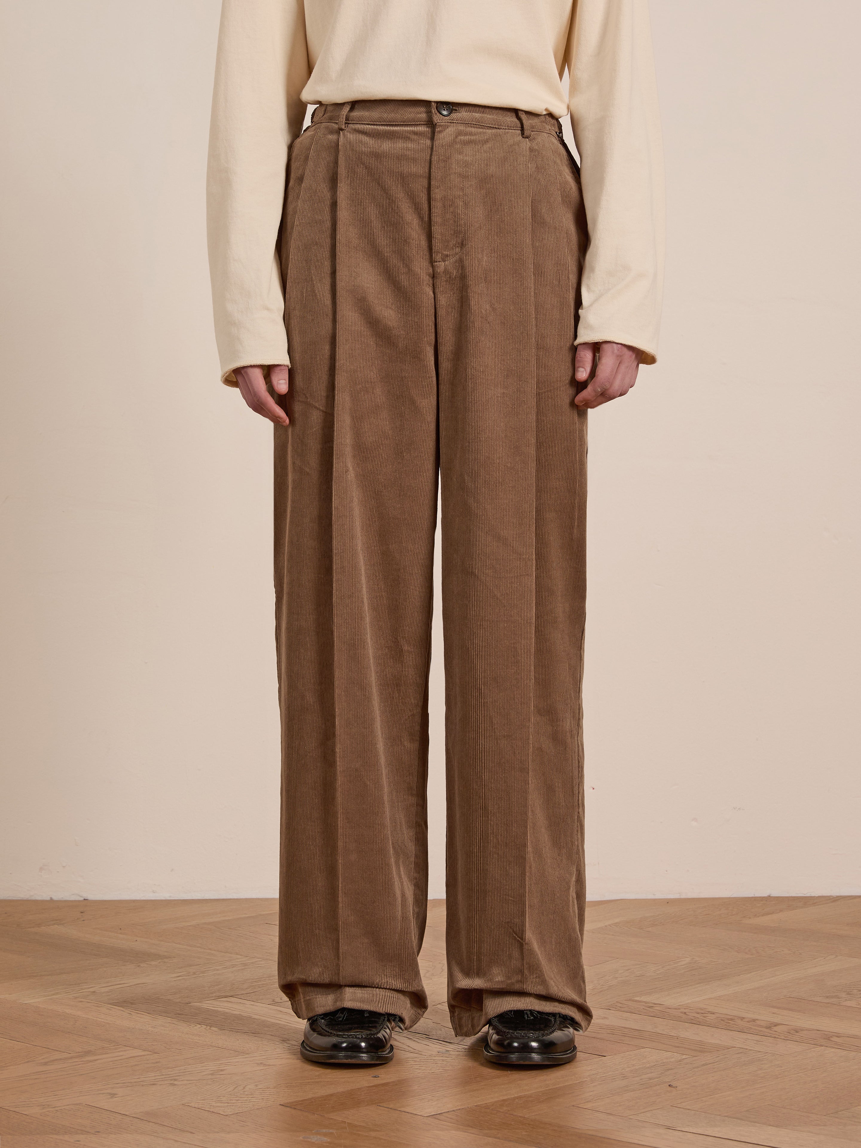 A person stands on a wooden floor wearing FOUND's Corduroy Pleated Trousers in brown, high-waisted and double-pleated, paired with a light beige long-sleeve shirt.