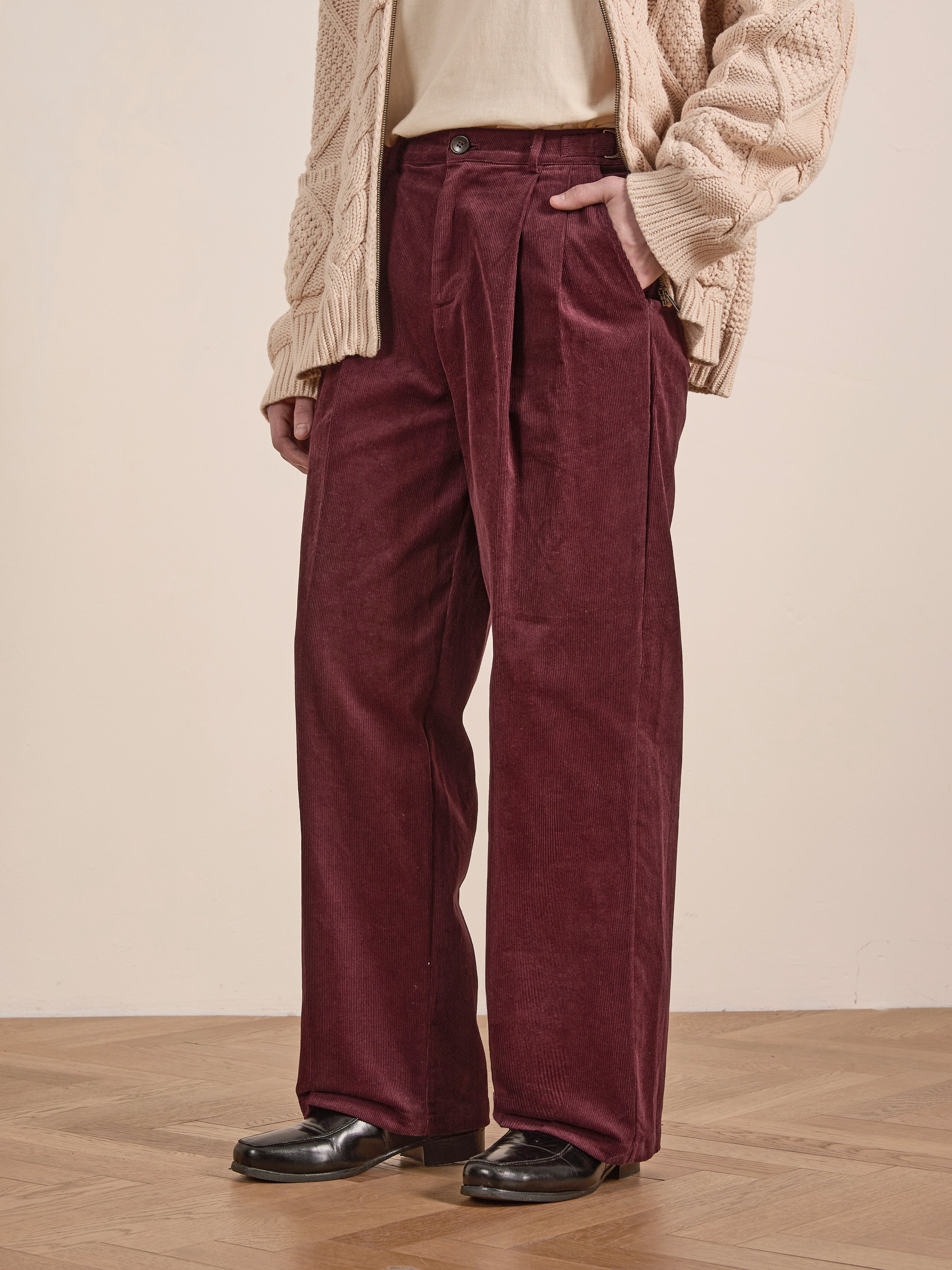 A person stands confidently on a wooden floor, wearing FOUND's unisex Corduroy Pleated Trouser in burgundy, paired with a cozy beige sweater and sleek black shoes.