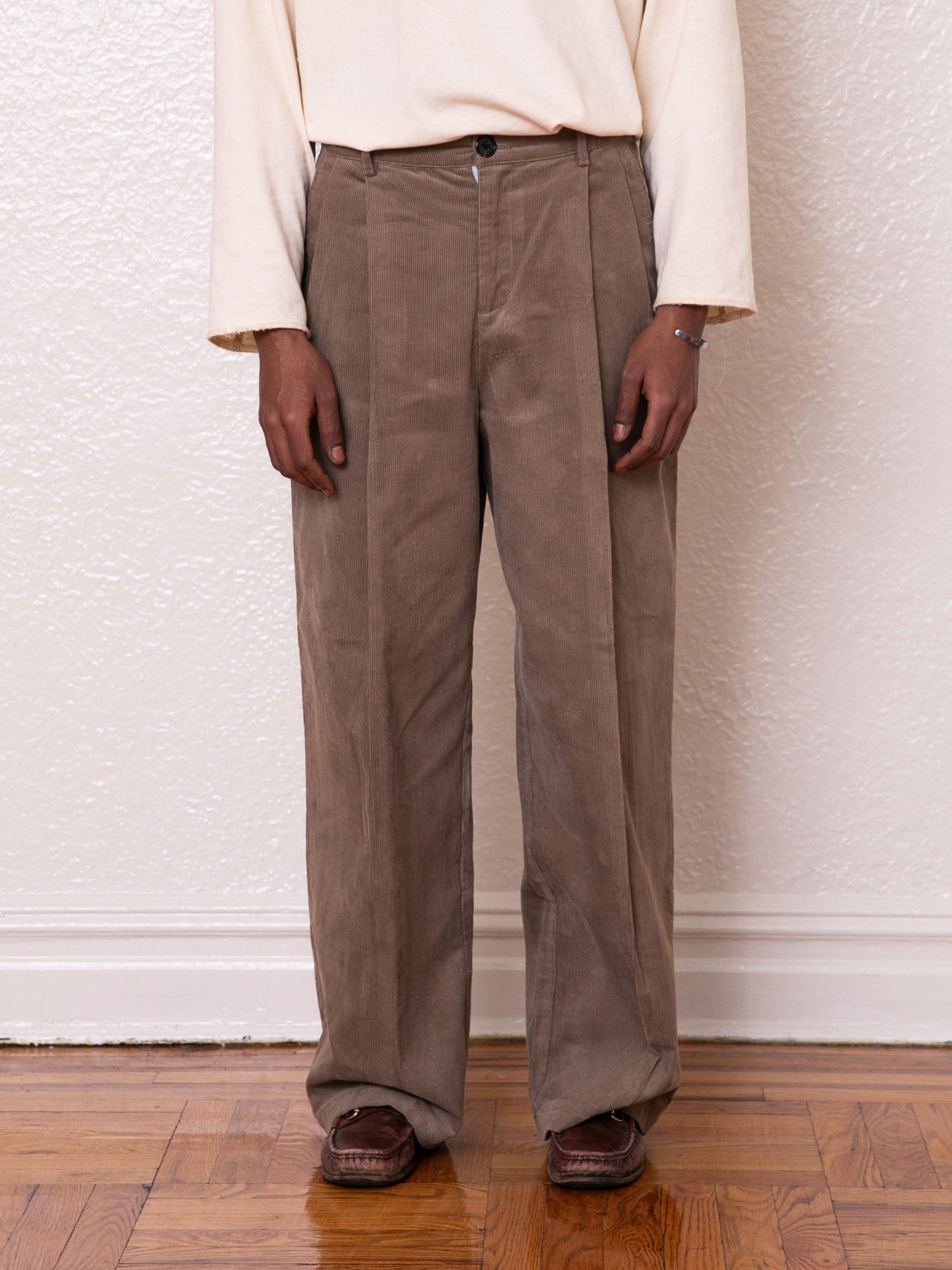 A person dressed in FOUND's high-rise Corduroy Pleated Trousers and a beige shirt stands in brown shoes on a wooden floor against a textured wall.