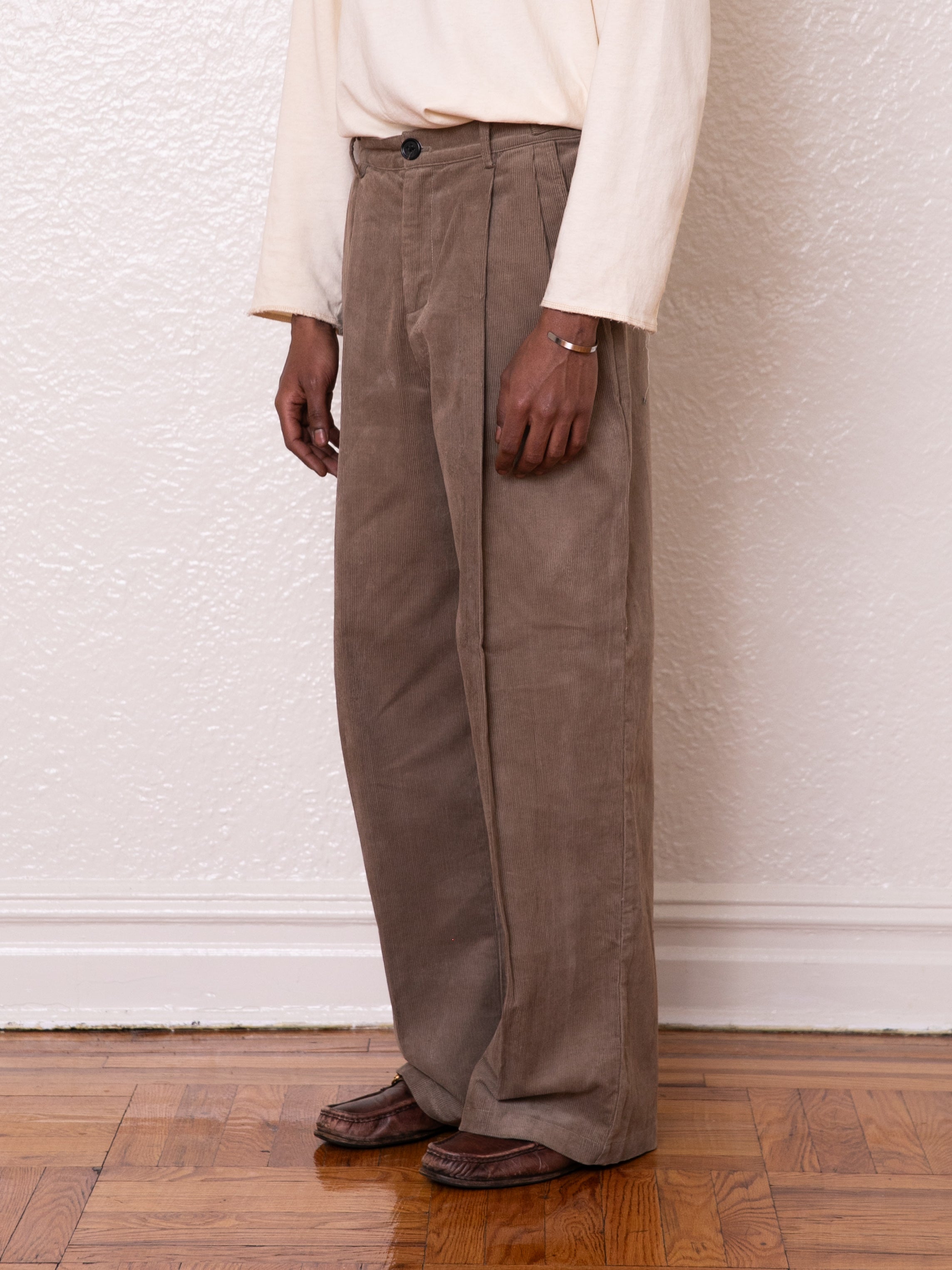 A person wearing a light long-sleeve shirt, FOUND's Corduroy Pleated Trousers that are brown with a high-rise waist, and matching shoes stands against a white textured wall on a wooden floor.