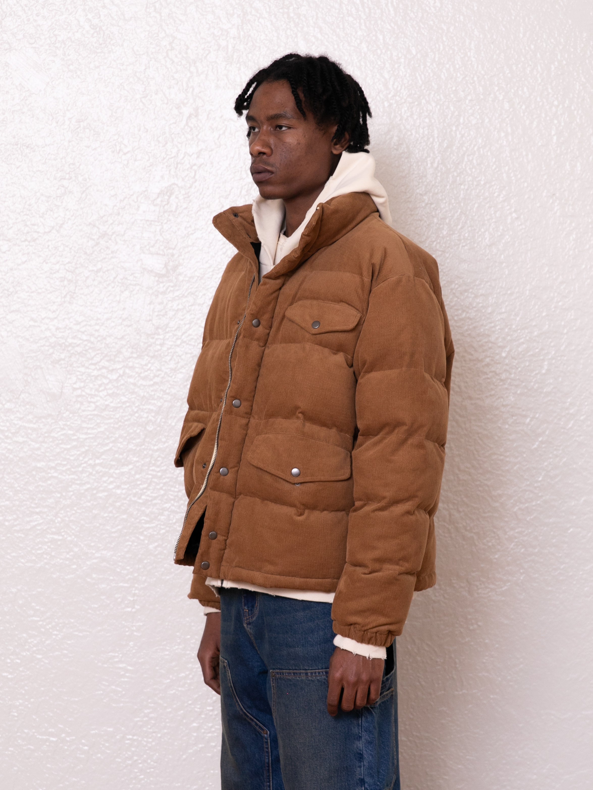 A person wearing a relaxed boxy fit Corduroy Puffer Jacket by FOUND in brown, layered over a white hoodie, stands against a textured white wall.