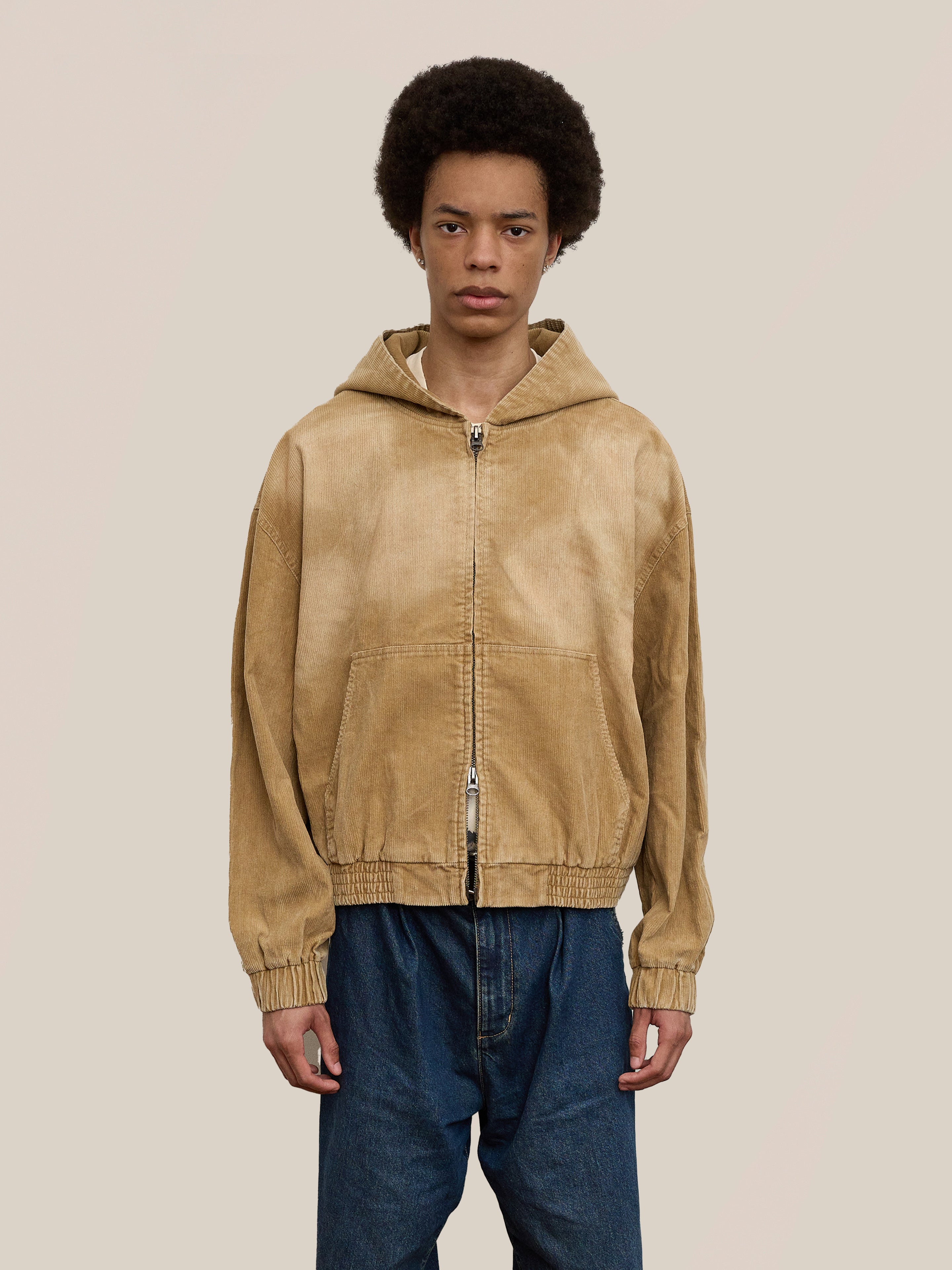A person wears the FOUND Corduroy Zip Up Hoodie in tan with blue jeans, standing facing forward against a beige background to showcase its unisex relaxed fit.