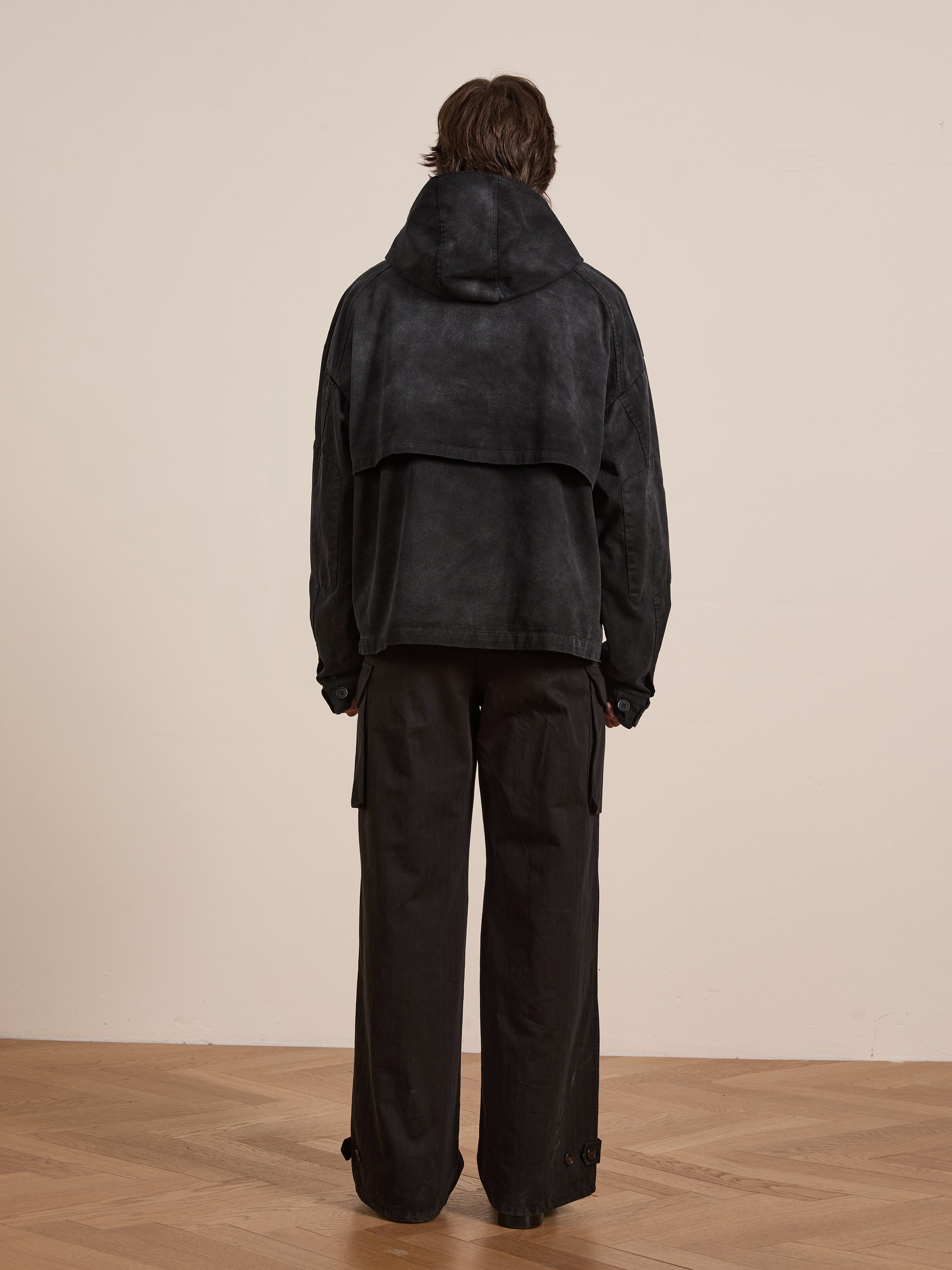 Clad in a FOUND Washed Twill Pocket Anorak Jacket and loose pants, the person stands on a wooden floor with their back to the camera, facing a plain wall.