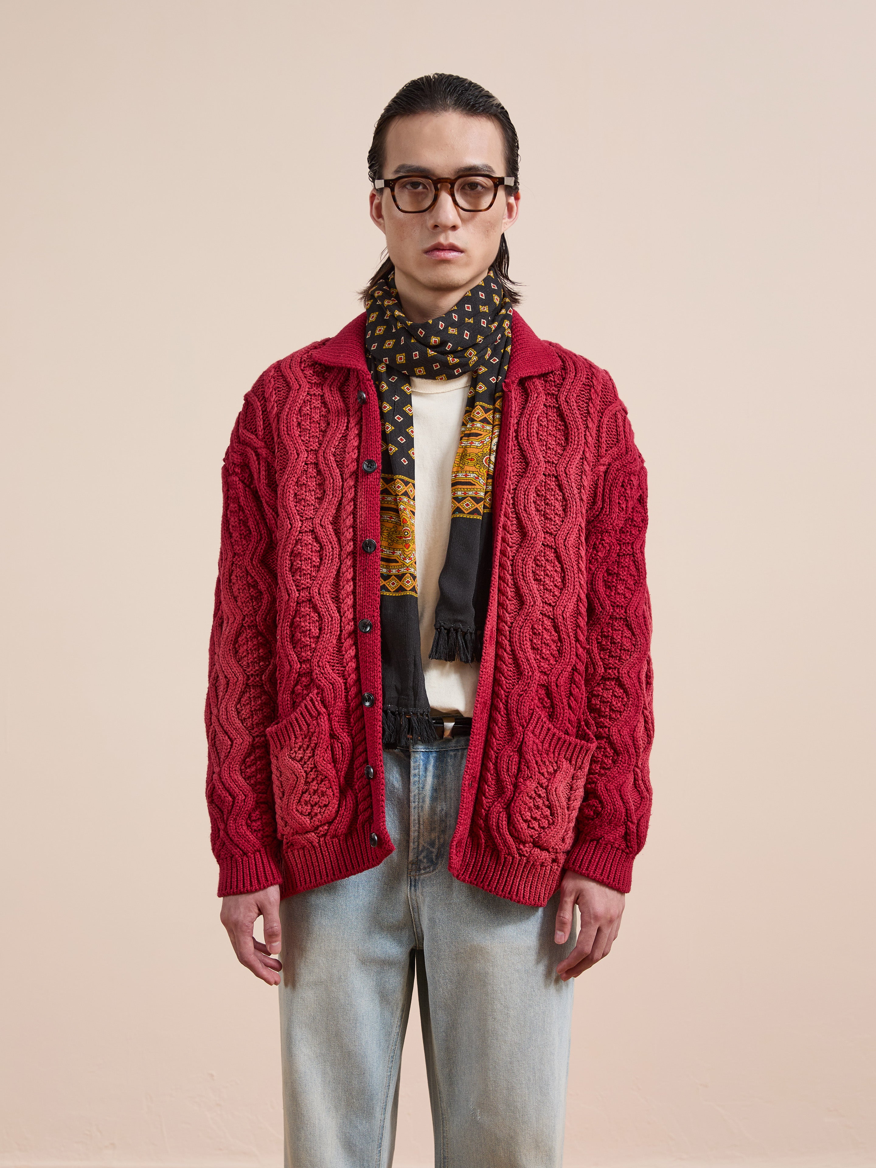 A person wearing glasses, the FOUND Cable Knit Buttoned Cardigan in red, a patterned scarf, white shirt, and light blue jeans stands facing the camera against a plain background.