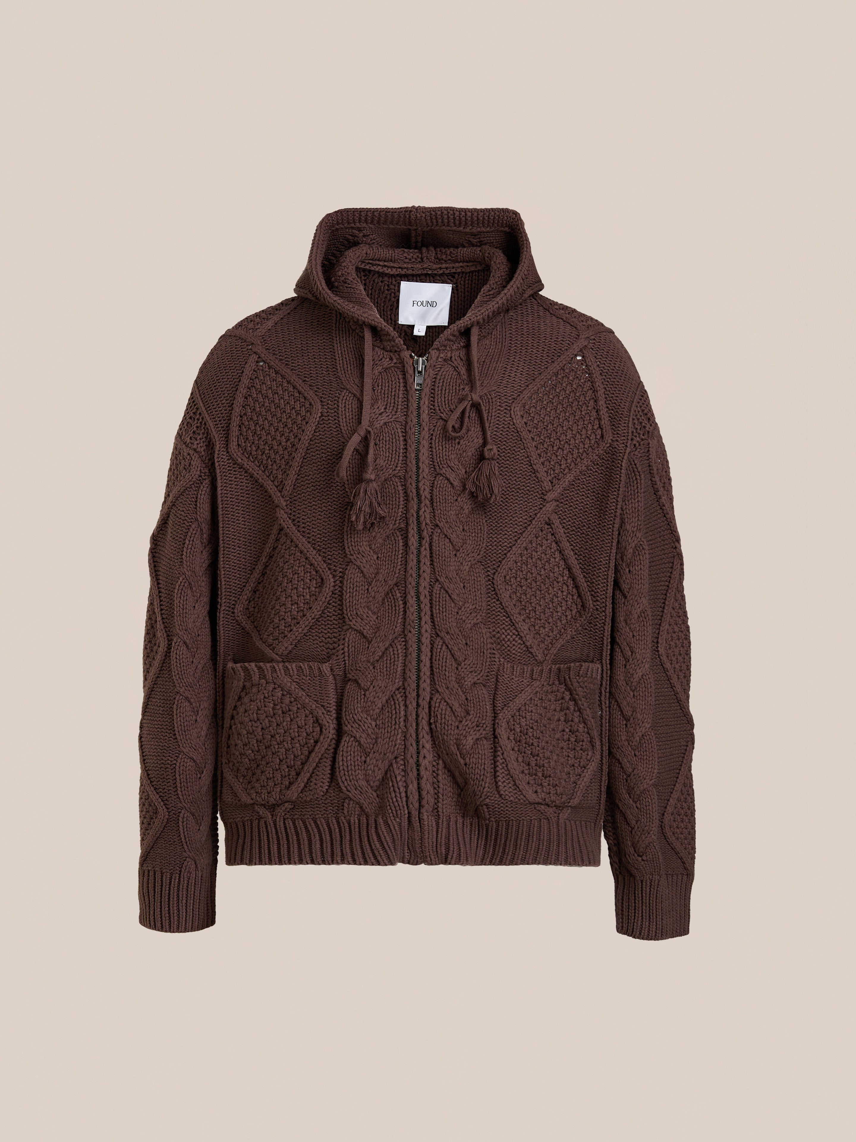 The FOUND Zip Up Cable Knit Hoodie features a unisex relaxed fit, front pockets, and hood. Vintage-inspired and comfortable, it’s shown against a plain beige background for timeless style.
