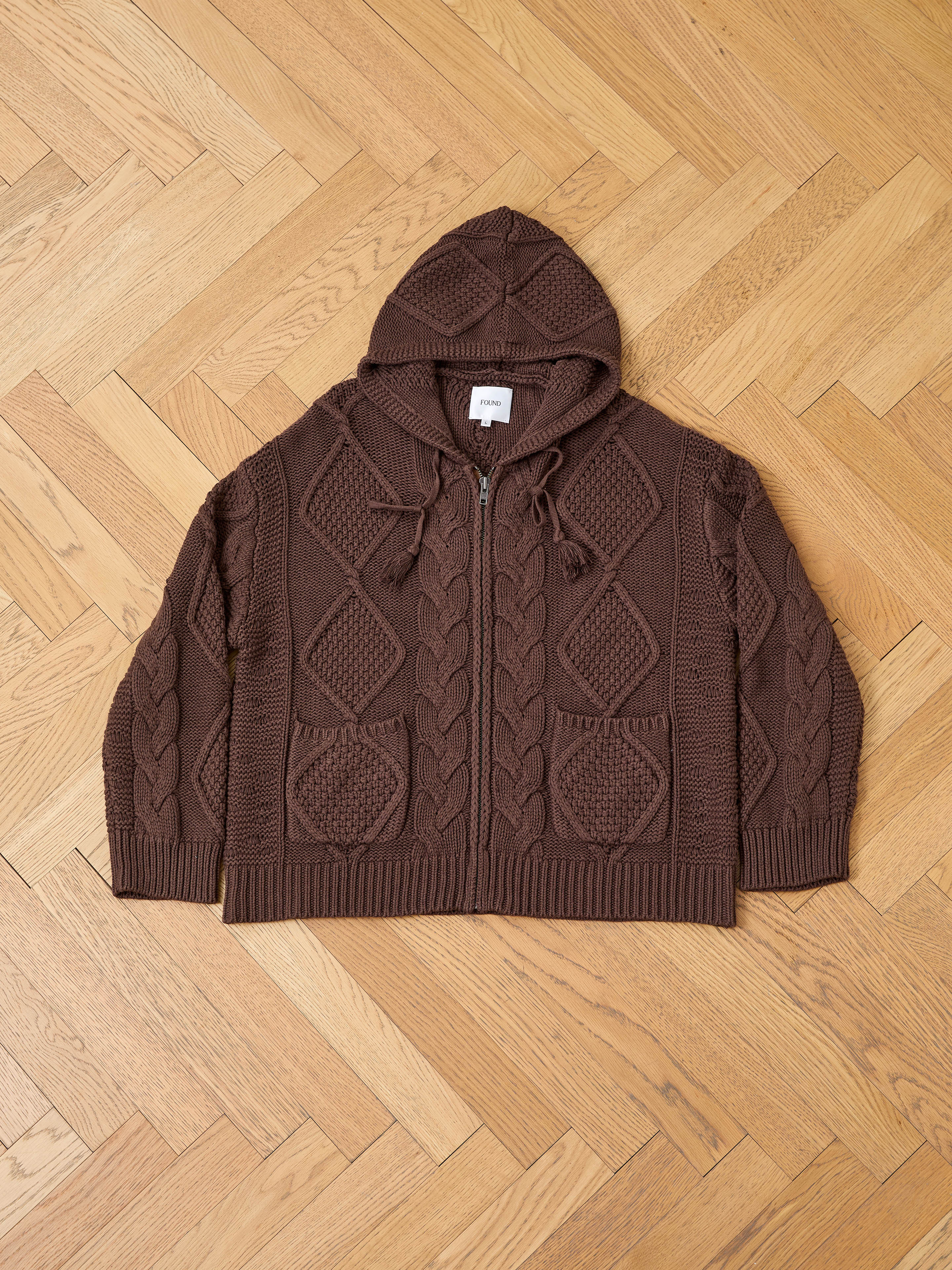 The FOUND Zip Up Cable Knit Hoodie features a front zipper, pockets, and a unisex relaxed fit, shown laid flat on a light wood herringbone floor.
