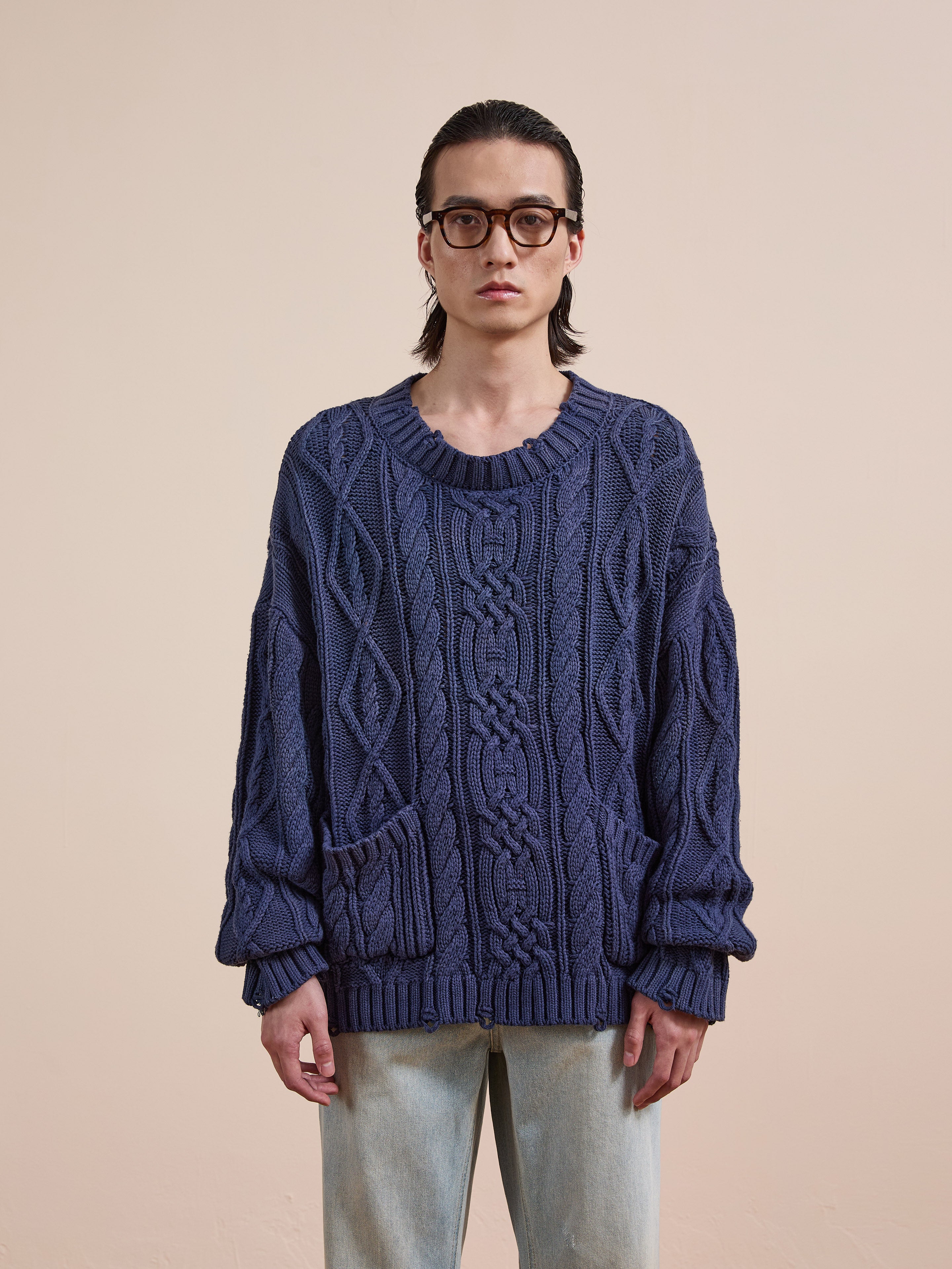 A person with long hair and glasses wears the Found Cable Knit Pullover Sweater, a loose-fitting unisex design with two front pockets, paired with light blue jeans, standing against a plain beige background.