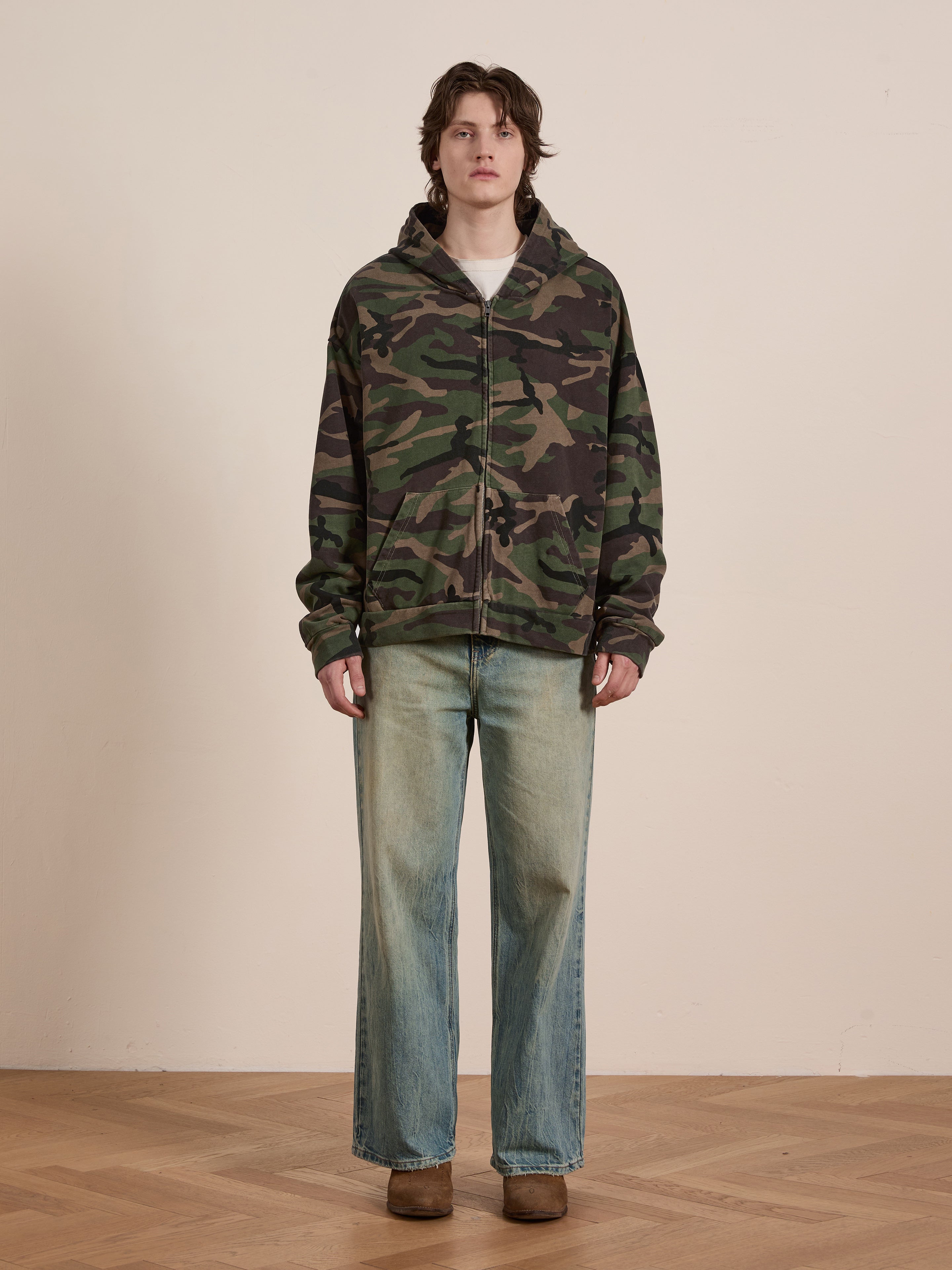 Against a plain wall, someone indoors wears the FOUND Washed Camouflage Zip Hoodie made from French terry cotton, paired with light blue wide-leg jeans and brown shoes.