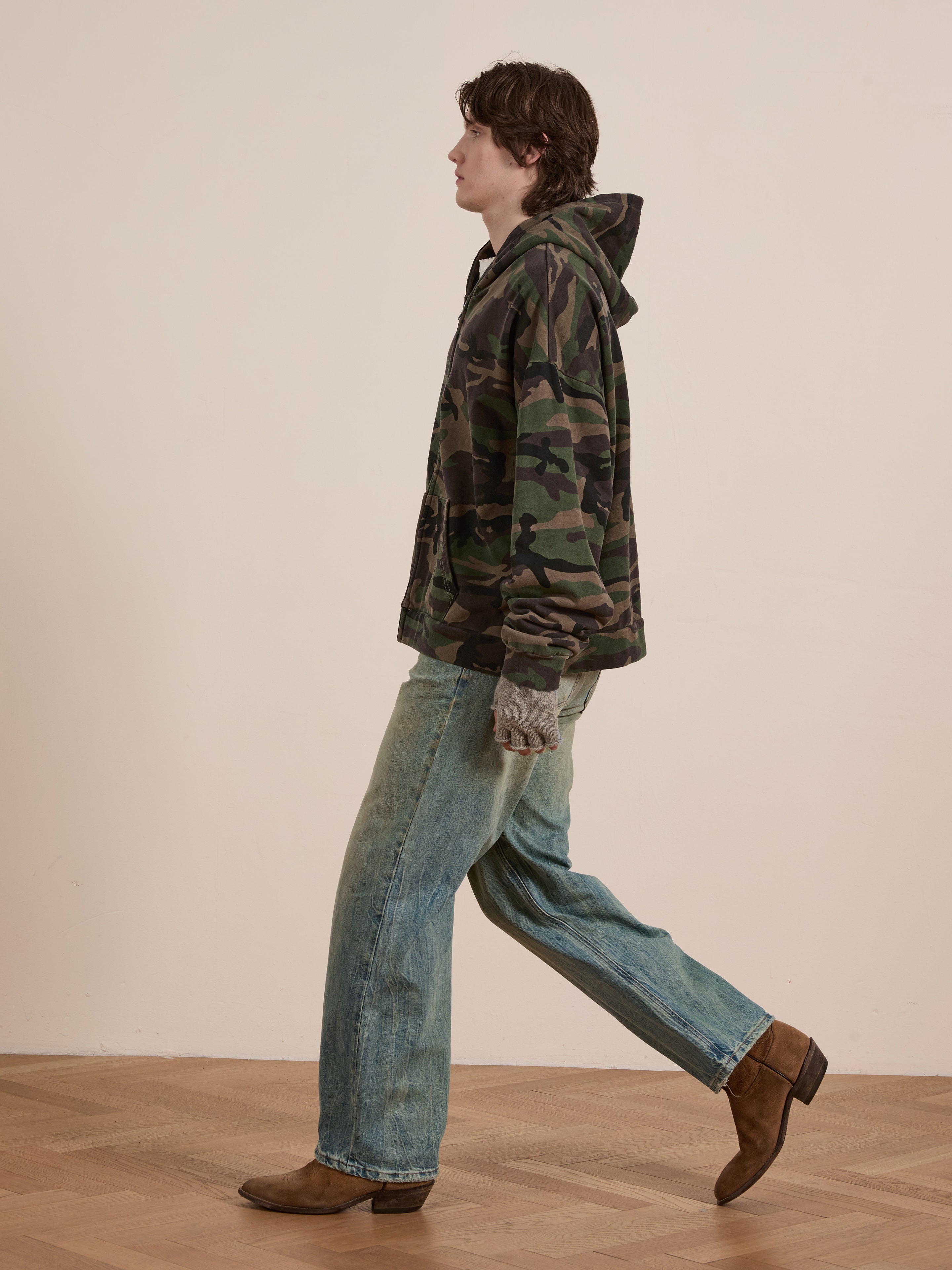 A side view of someone in the FOUND Washed Camouflage Zip Hoodie, jeans, and brown boots walking indoors on a wooden floor.