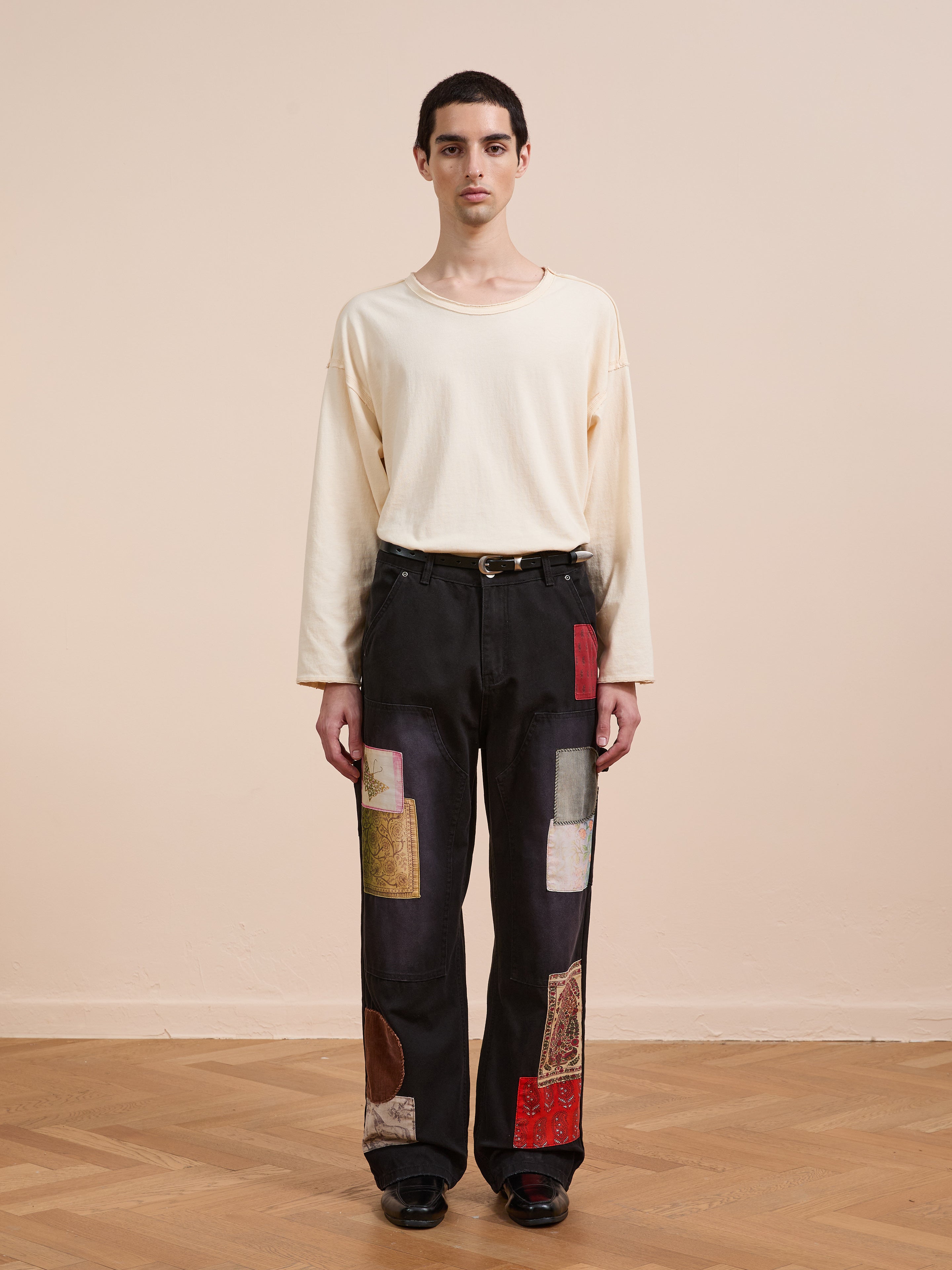 A person stands indoors facing the camera, wearing FOUND Tapestry Carpenter Canvas Pants featuring a relaxed fit and Pakistani quilt-inspired prints, paired with a loose cream long-sleeve shirt against a plain beige background.