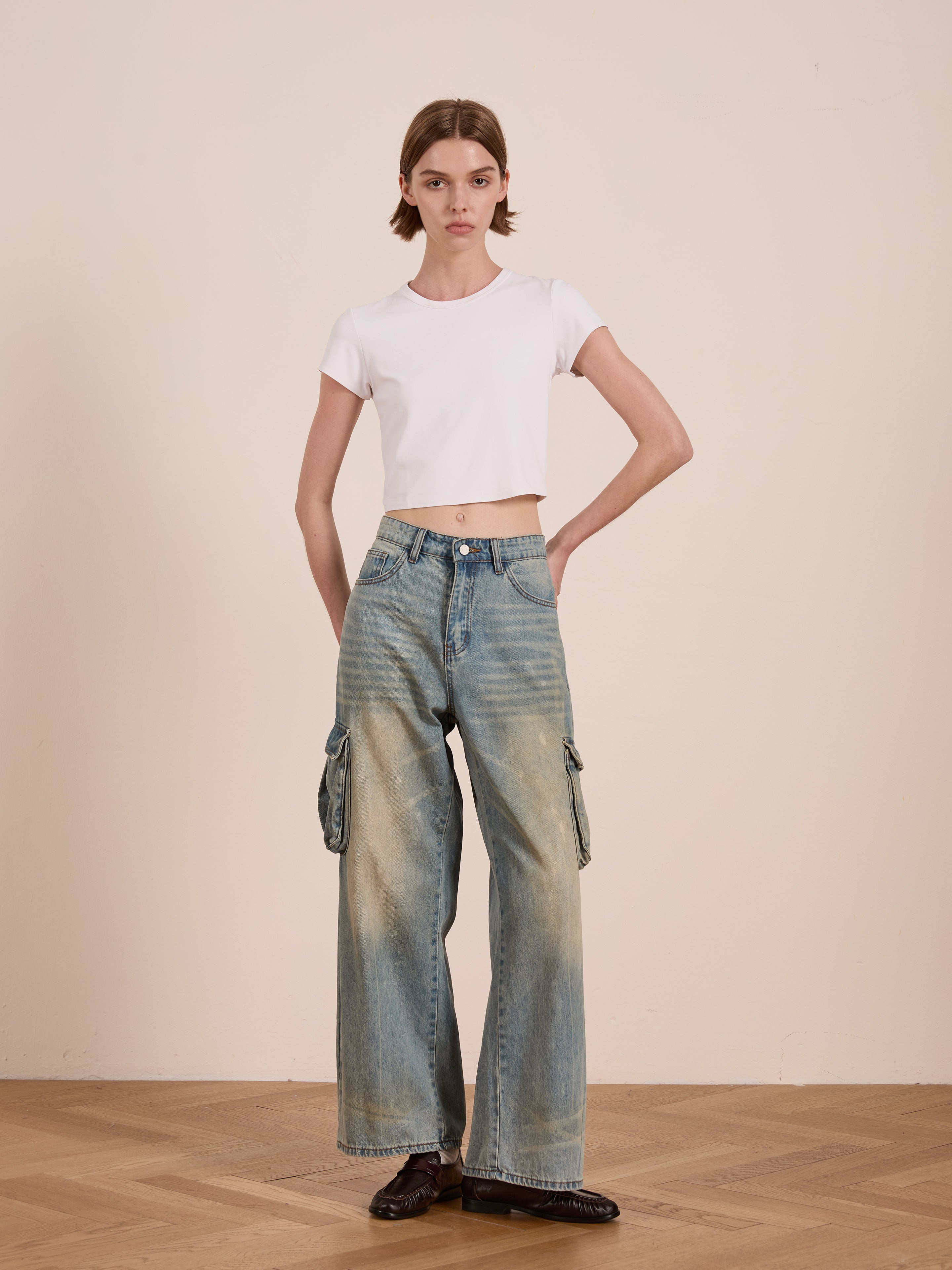 A person sporting a white crop top and FOUND's Cargo Jeans, featuring a relaxed fit and utilitarian edge, stands on a wooden floor against a plain wall.