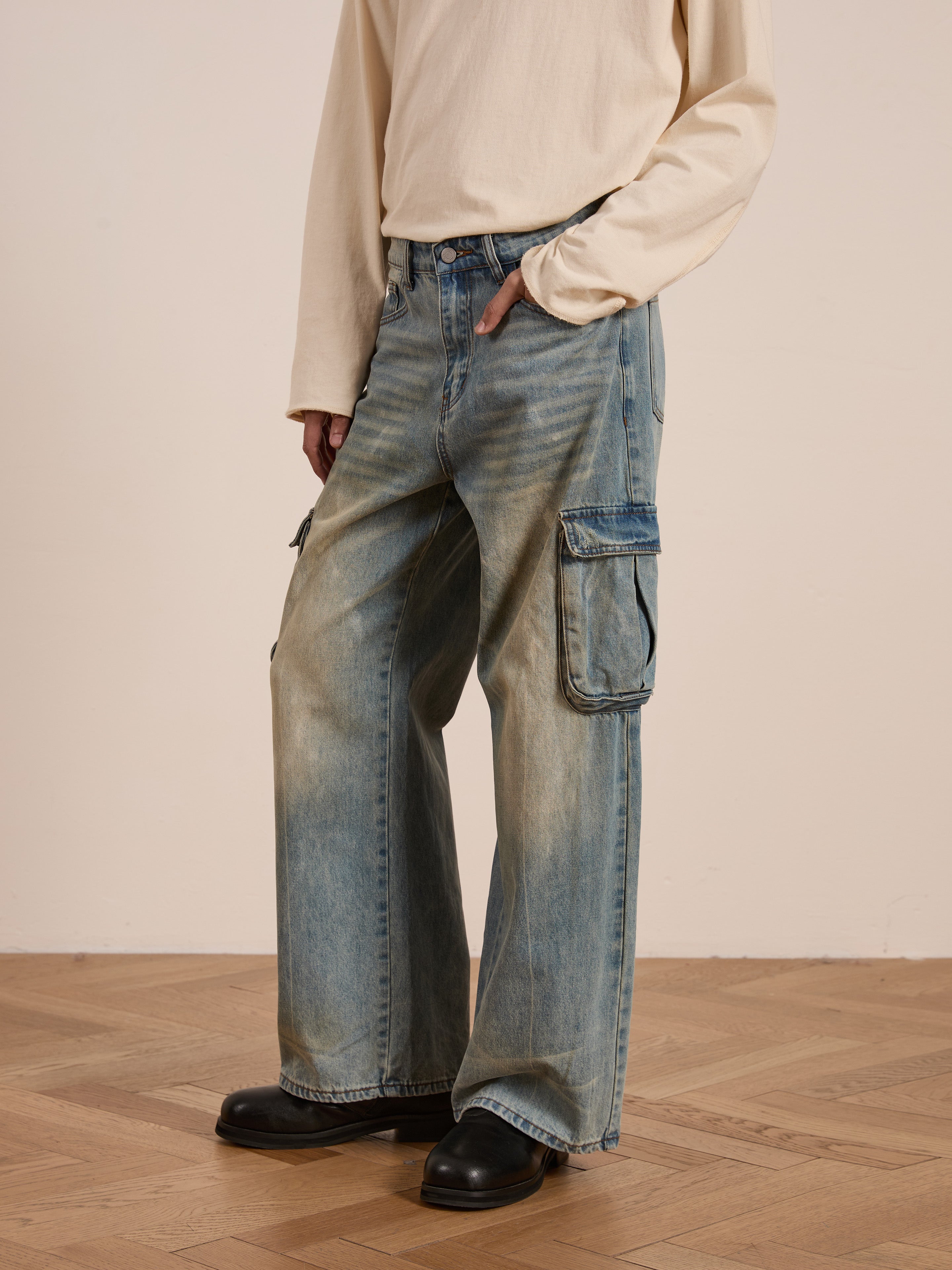 Standing on a wooden floor, a person exudes utilitarian style wearing relaxed-fit FOUND Cargo Jeans with spacious pockets, paired with a long-sleeve beige shirt.