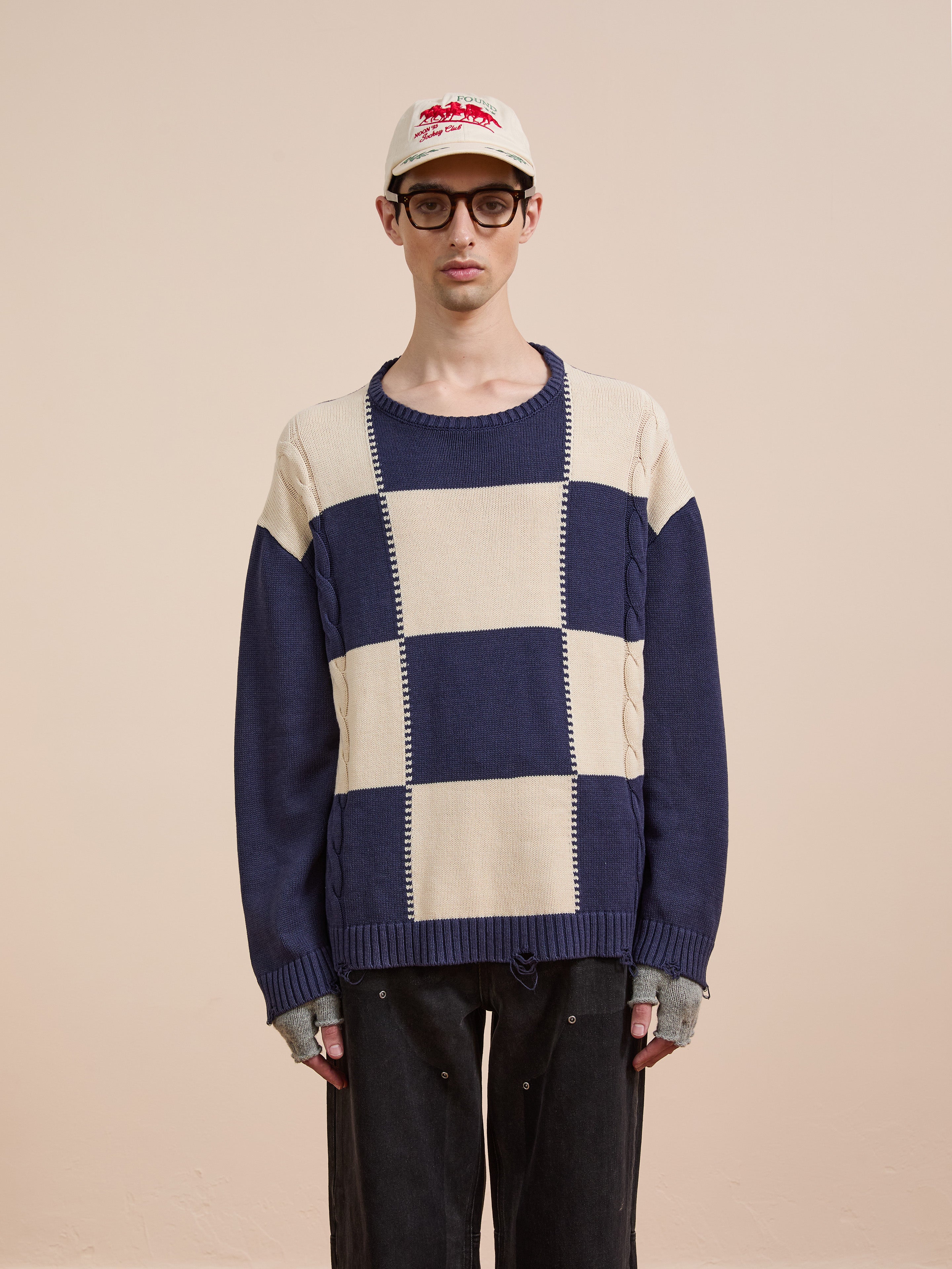 A person stands against a plain background wearing the Found Checkered Fisherman Sweater in cream and navy, paired with black pants, fingerless gloves, glasses, and a white cap.