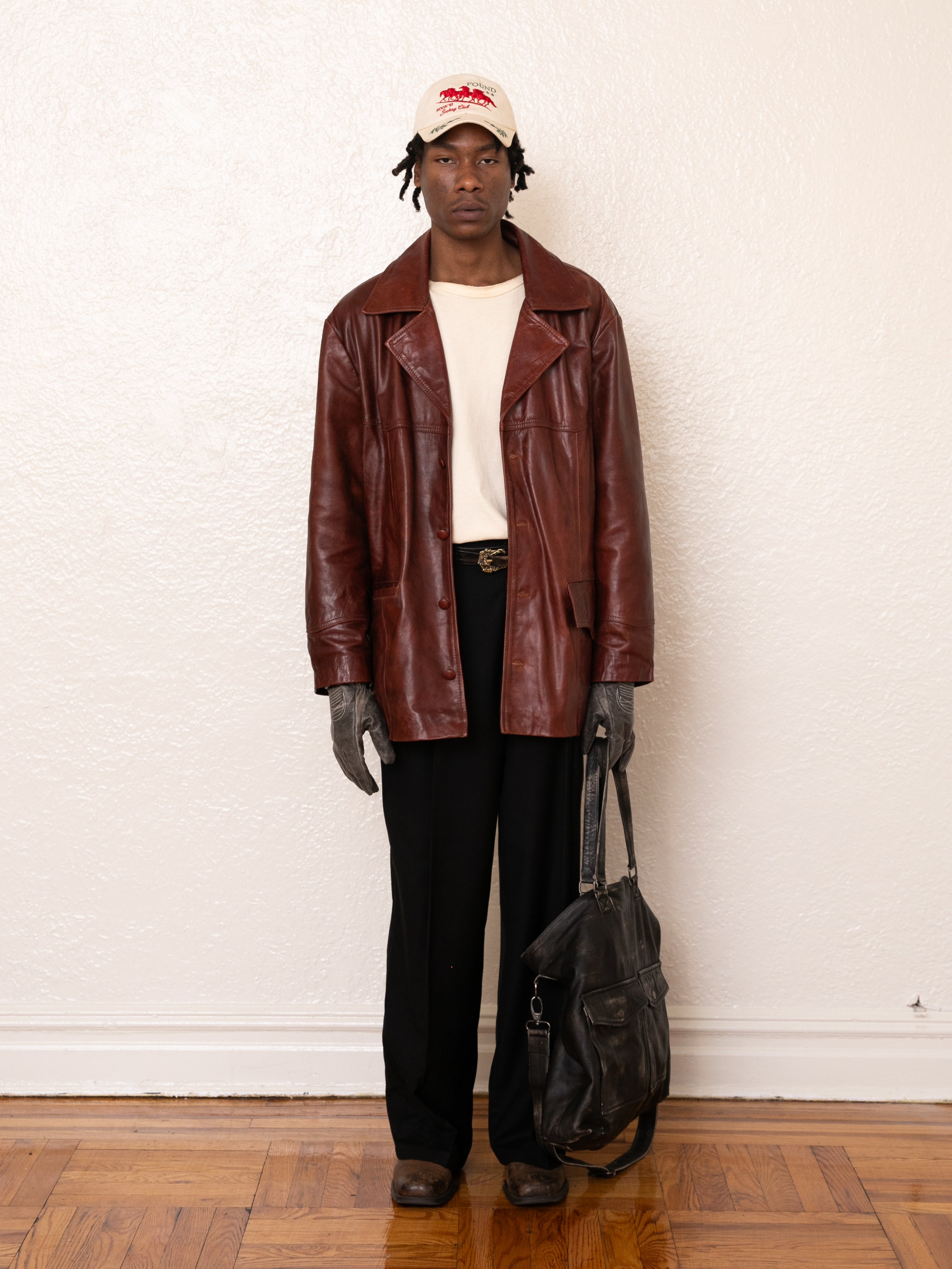 A person stands against a white wall wearing a Claret Western Leather Blazer from FOUND, paired with a cream shirt, black pants, gloves, and a white cap. They hold an ethically made black bag on the wooden floor.