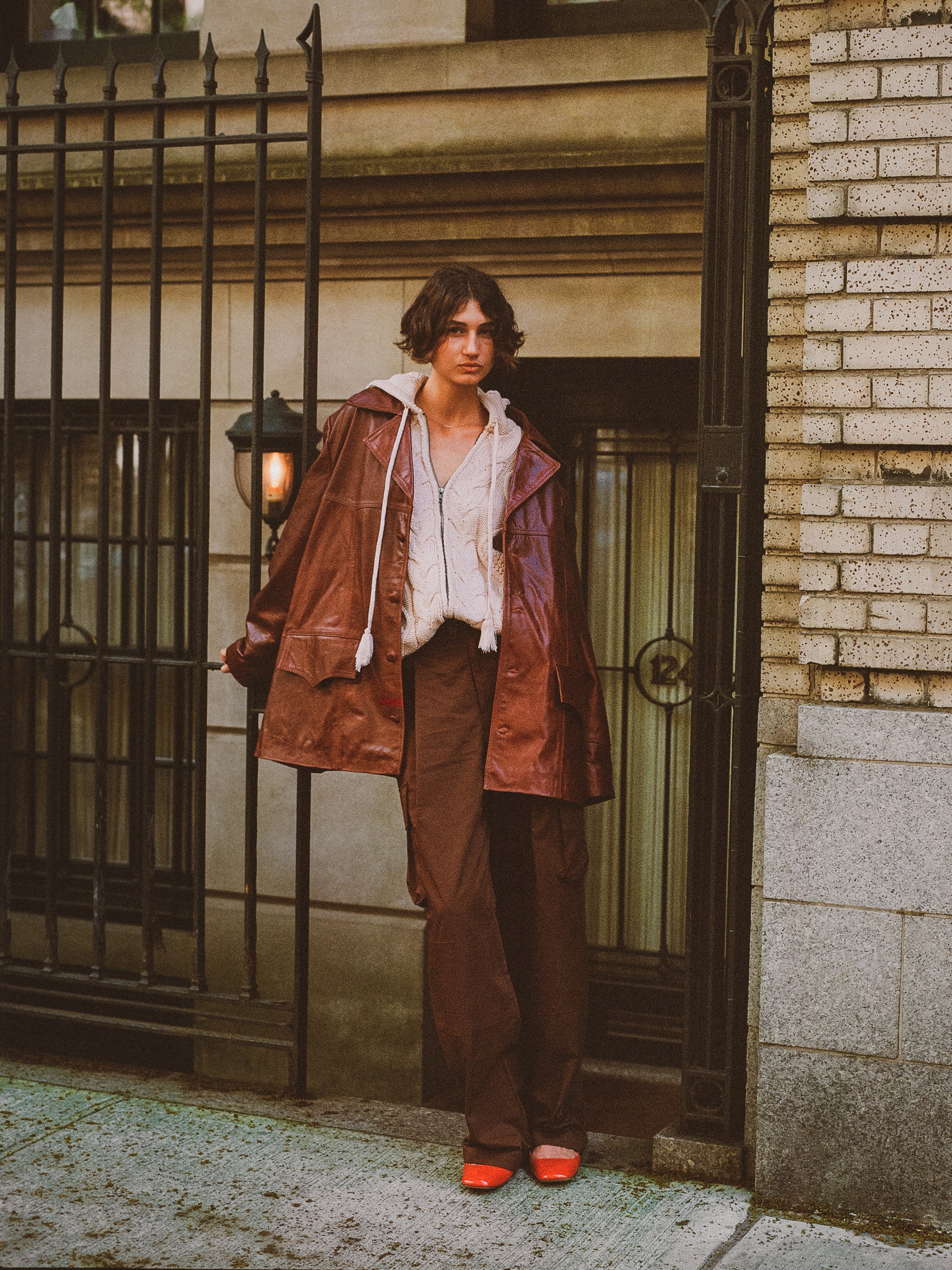 A person wearing a FOUND Claret Western Leather Blazer with a white shirt, brown pants, and red shoes stands by a brick building, holding the blazer open to showcase its lightweight cowhide material.