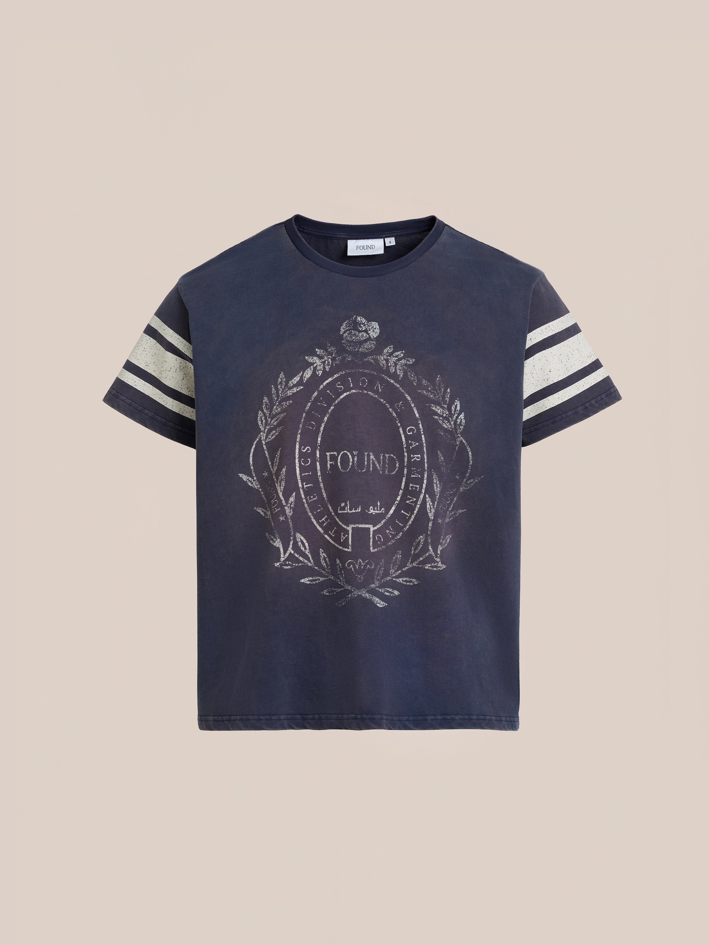 The Collegiate Crest Tee by FOUND is a navy blue unisex short-sleeve T-shirt with two white stripes on each sleeve and a decorative collegiate crest emblem with text on the front.