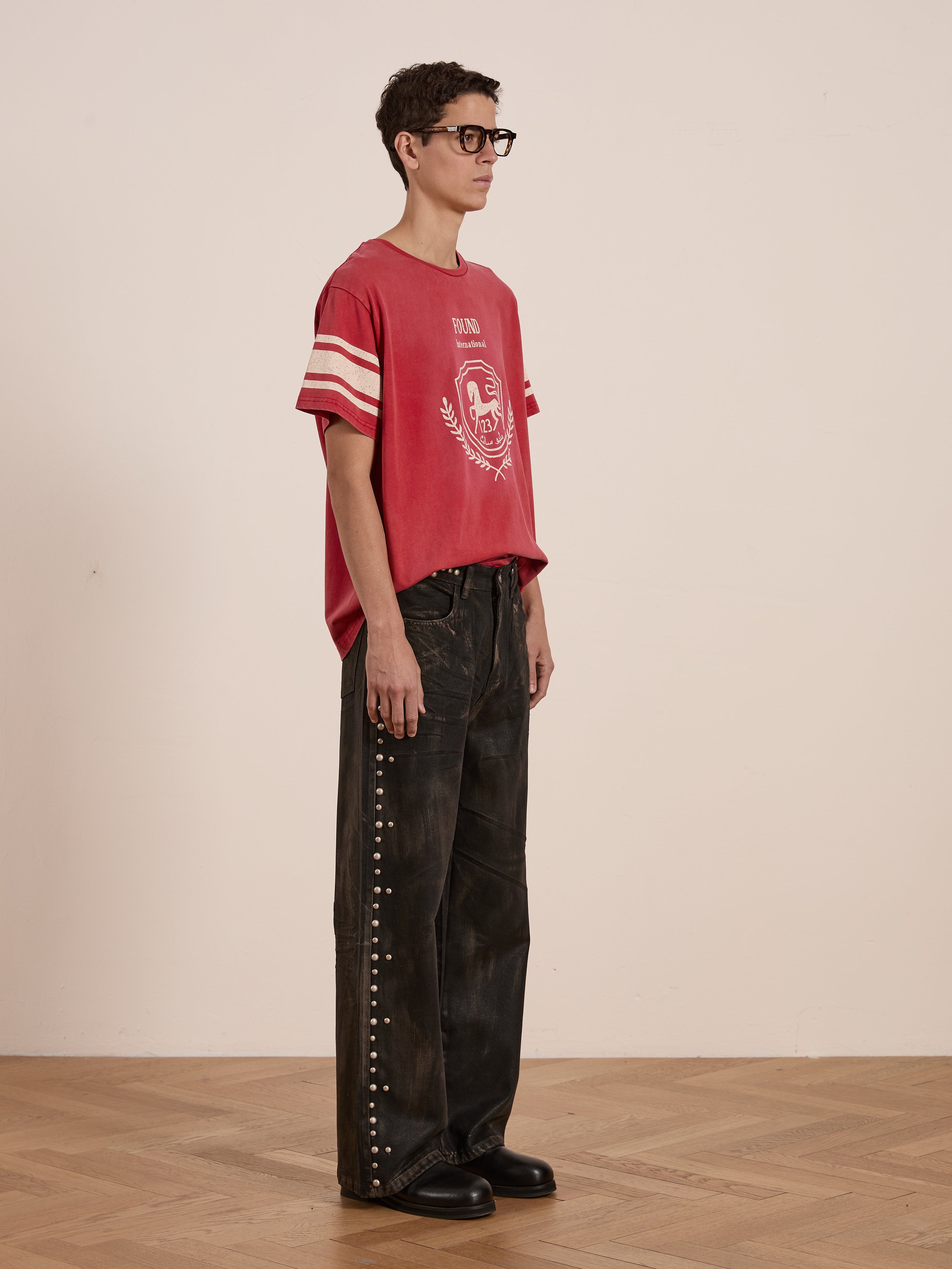 Person in a FOUND Collegiate Crest Tee, featuring a red hue and unisex relaxed fit, paired with black wide-leg pants with stud details, standing on a wooden floor indoors.