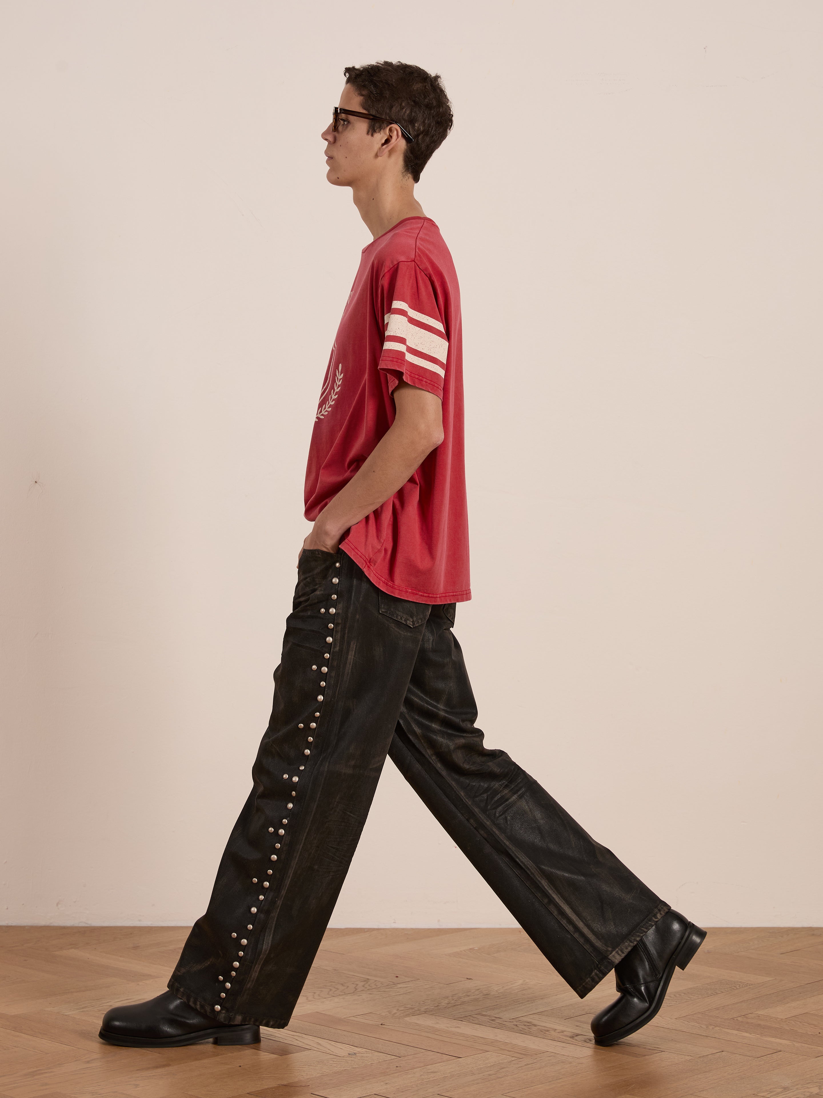 Wearing a FOUND Collegiate Crest Tee and studded black pants, a person strolls on a wooden floor, exuding the casual charm of vintage athletic gear against a plain backdrop.