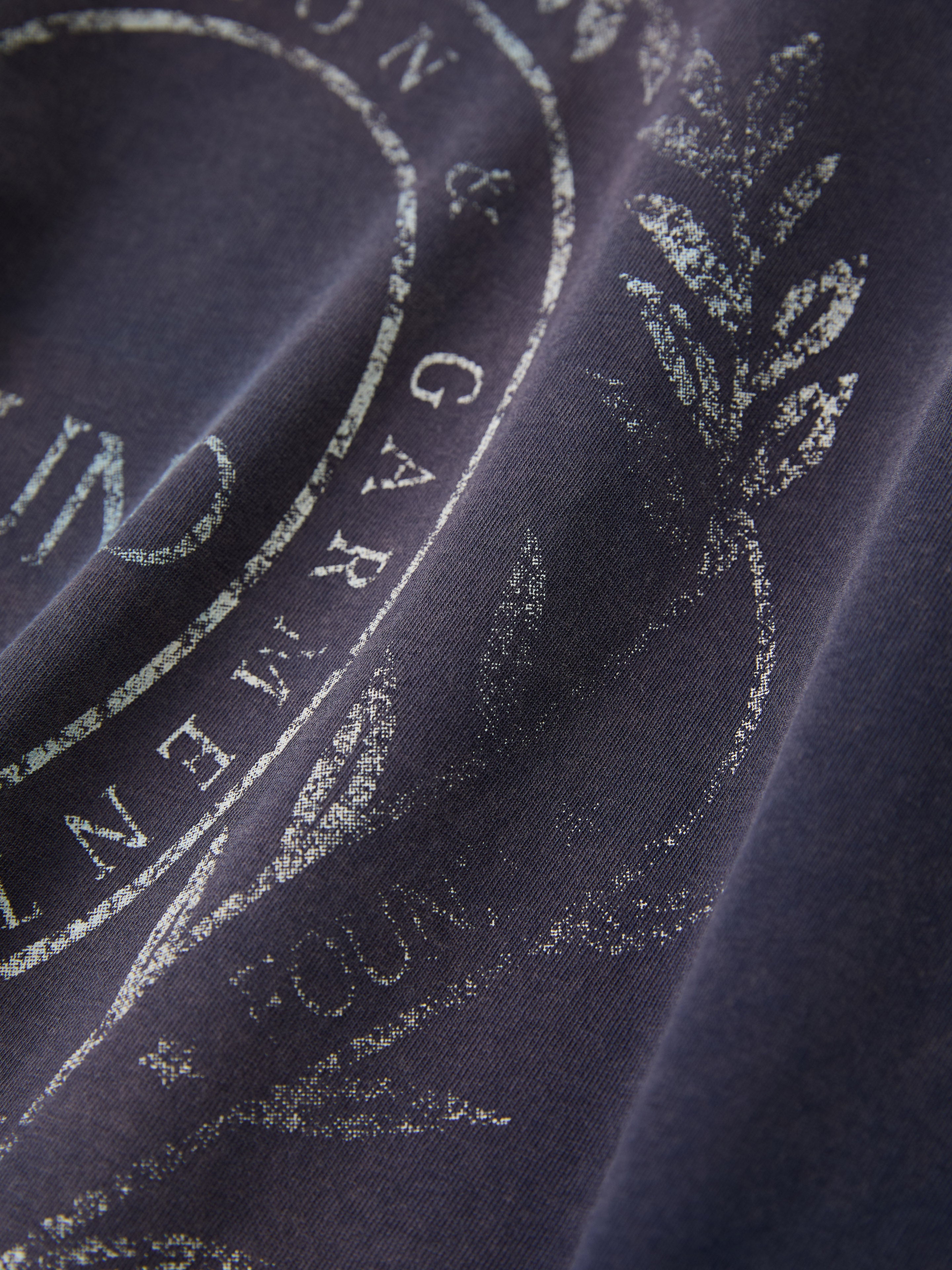 Close-up of a FOUND Collegiate Crest Tee, showcasing vintage athletic fabric in navy blue with a faded white circular insignia, decorative text, and laurel branch design.