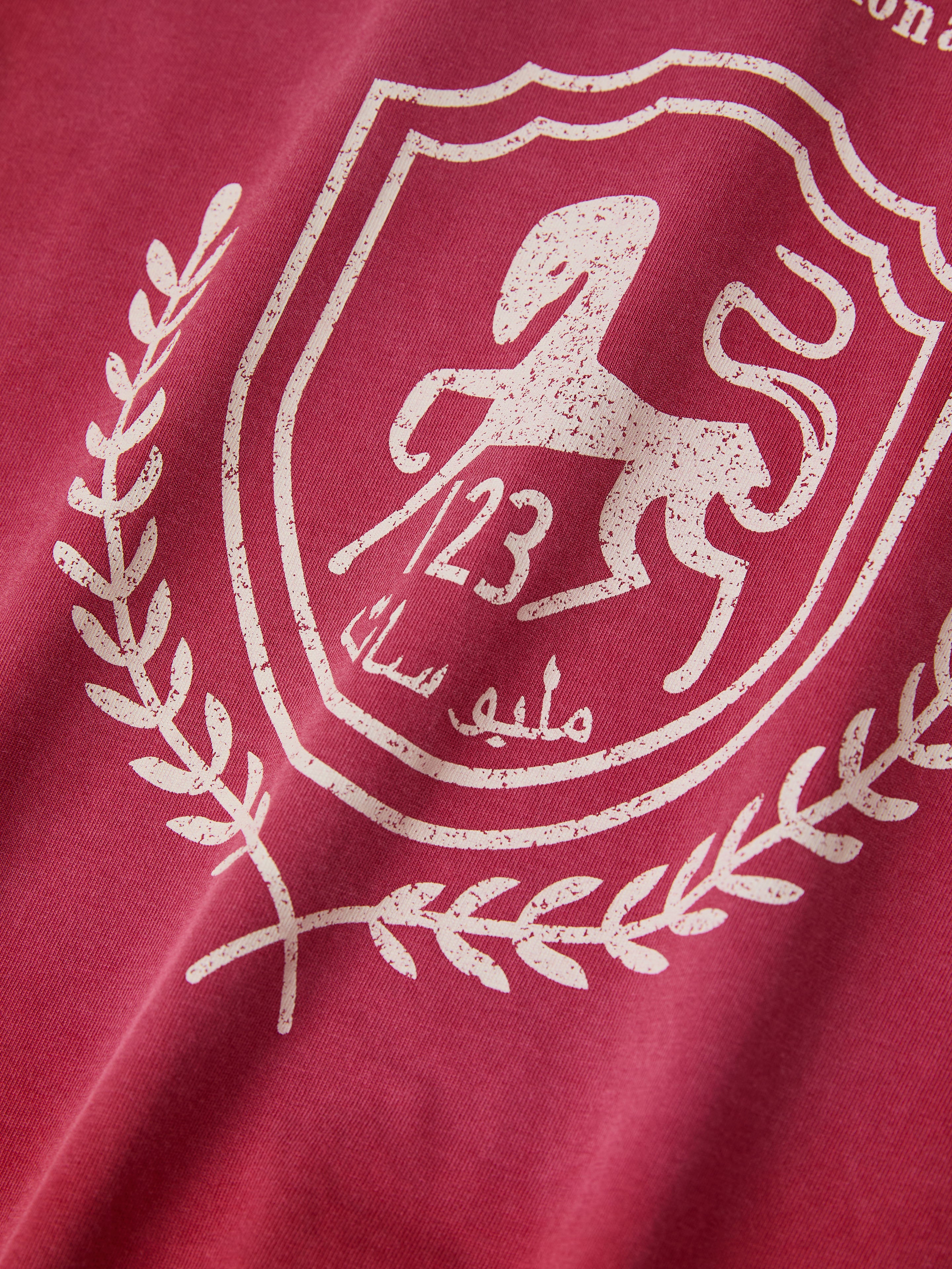 The red Collegiate Crest Tee by FOUND exudes nostalgic charm, featuring a white emblem with a stylized horse, laurel wreath, and number 123. Arabic text elegantly adorns this vintage athletic gear-inspired design.