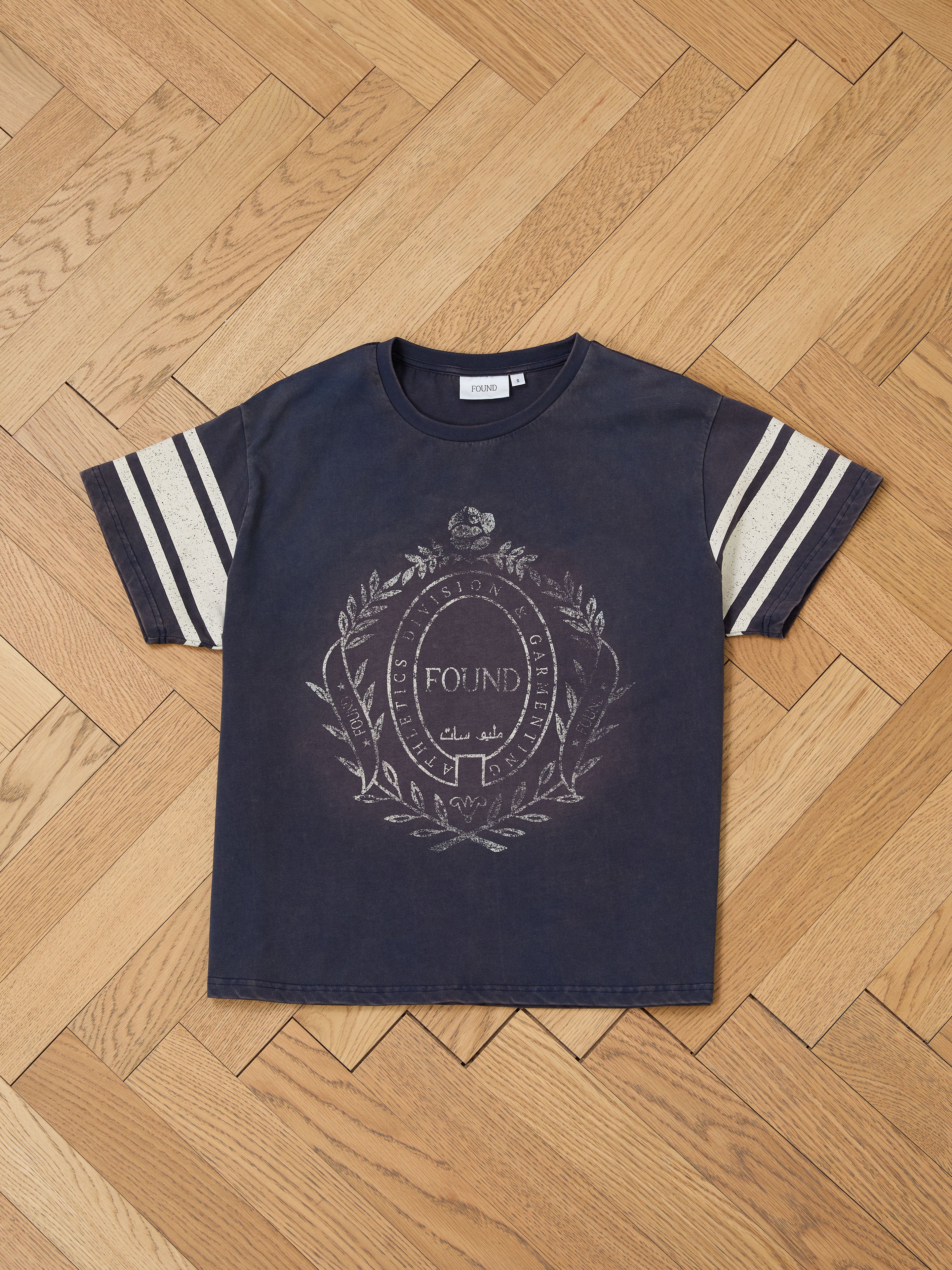 Laid elegantly on a herringbone floor, the FOUND Collegiate Crest Tee is a vintage unisex piece featuring a dark gray color, white sleeve stripes, and a classic collegiate design.