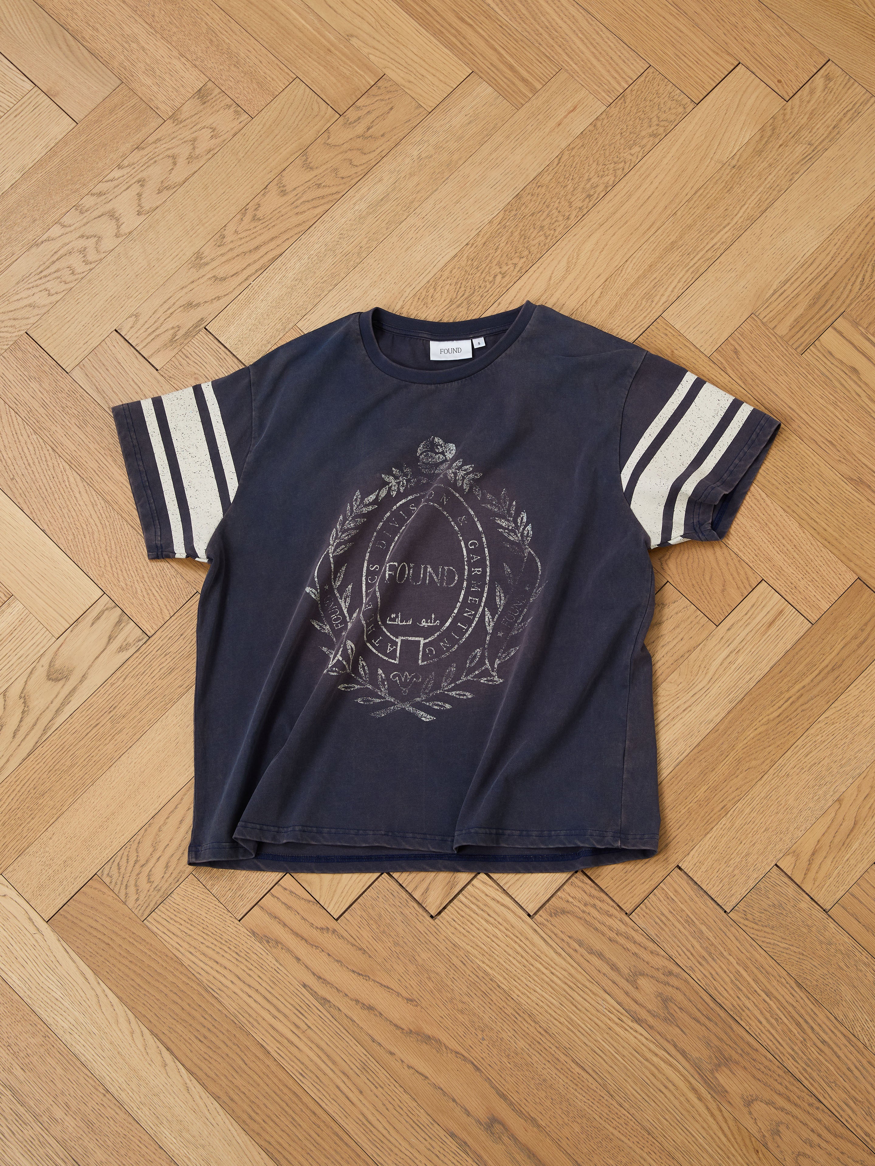 The FOUND Collegiate Crest Tee, featuring a faded black color, striped short sleeves, and a front emblem reminiscent of vintage athletic wear, rests on a wooden herringbone floor. This unisex design fuses classic style with modern appeal.