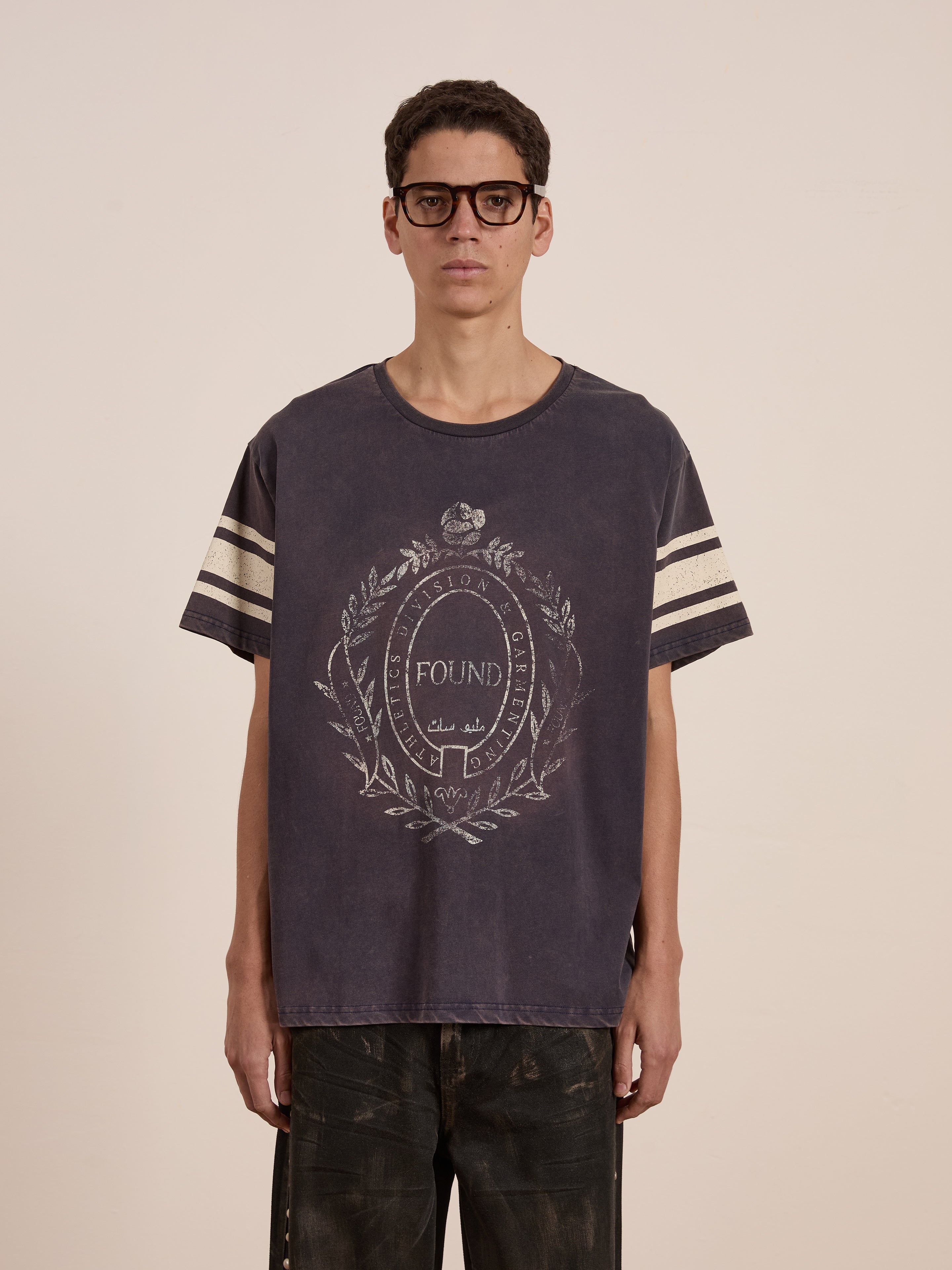 A person wearing glasses and a dark, distressed FOUND Collegiate Crest Tee with striped sleeves and a circular vintage athletic design stands against a plain background. This unisex shirt embodies the timeless appeal of a collegiate crest tee.