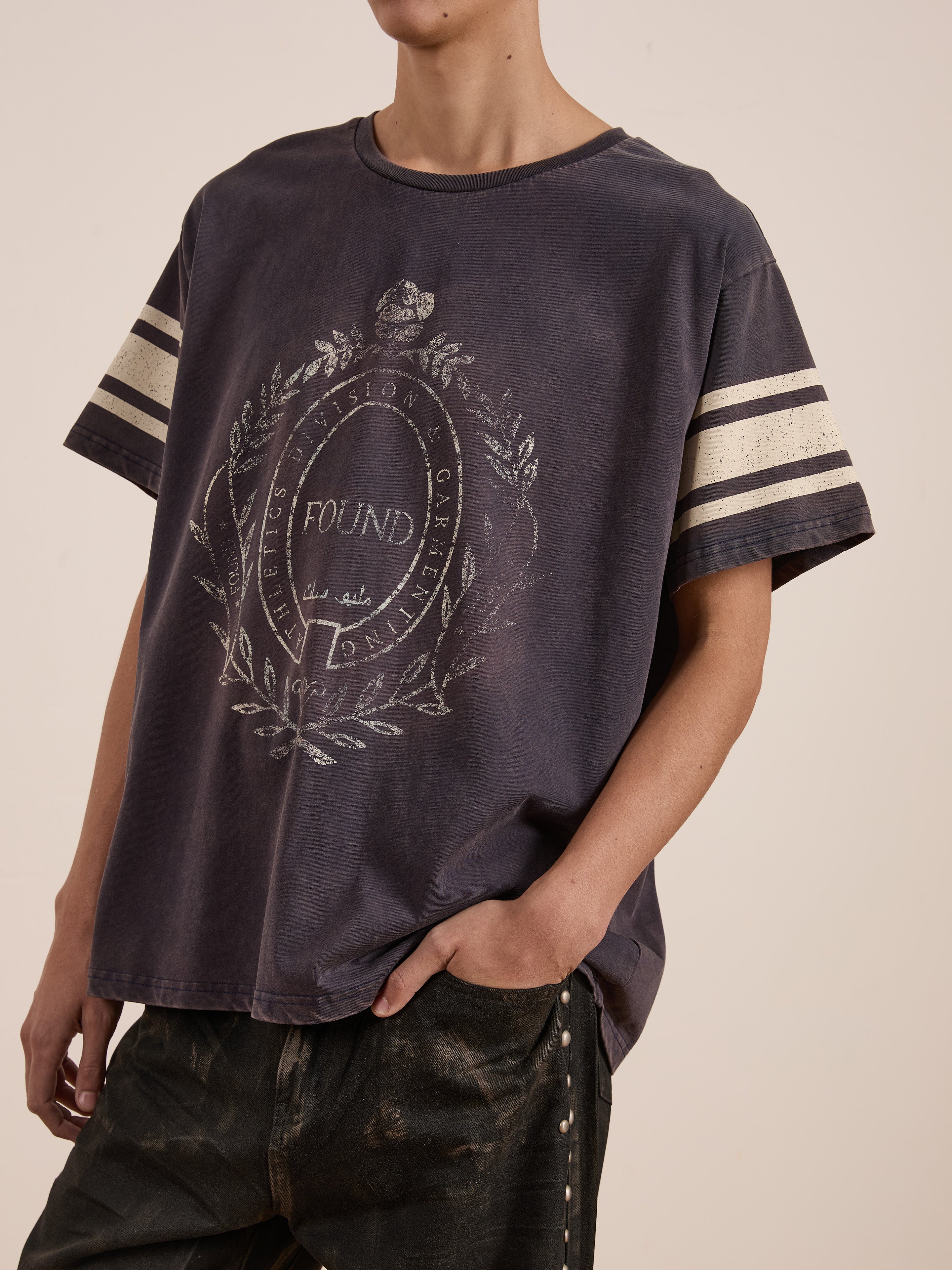 A person in a dark, oversized FOUND Collegiate Crest Tee poses with one hand in a pocket, paired with dark pants for a vintage athletic style.