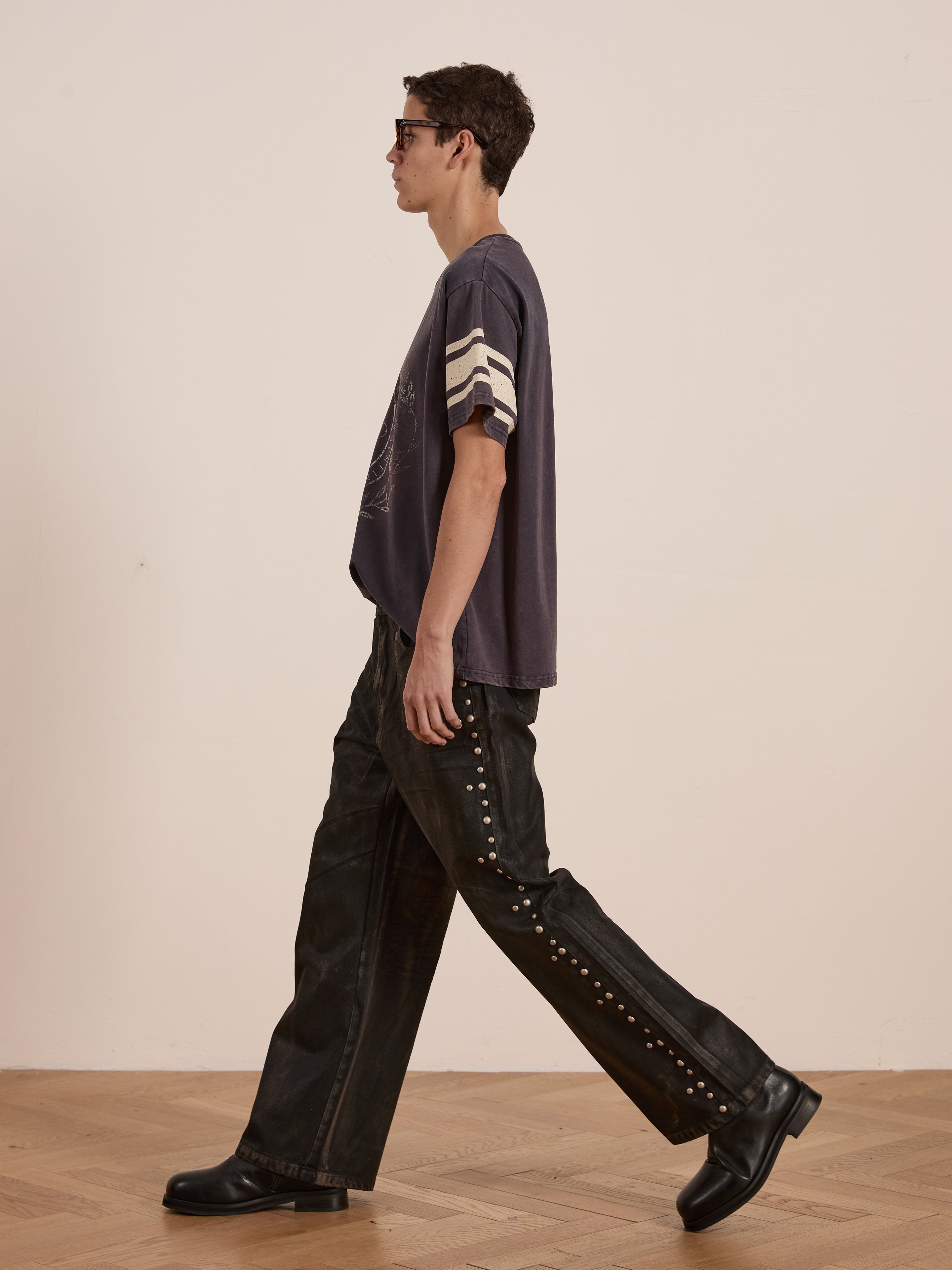 A person wearing FOUND's Collegiate Crest Tee and sunglasses, paired with dark pants featuring side embellishments and black shoes, strides across a wooden floor, exuding a unisex vibe that echoes vintage athletic style.