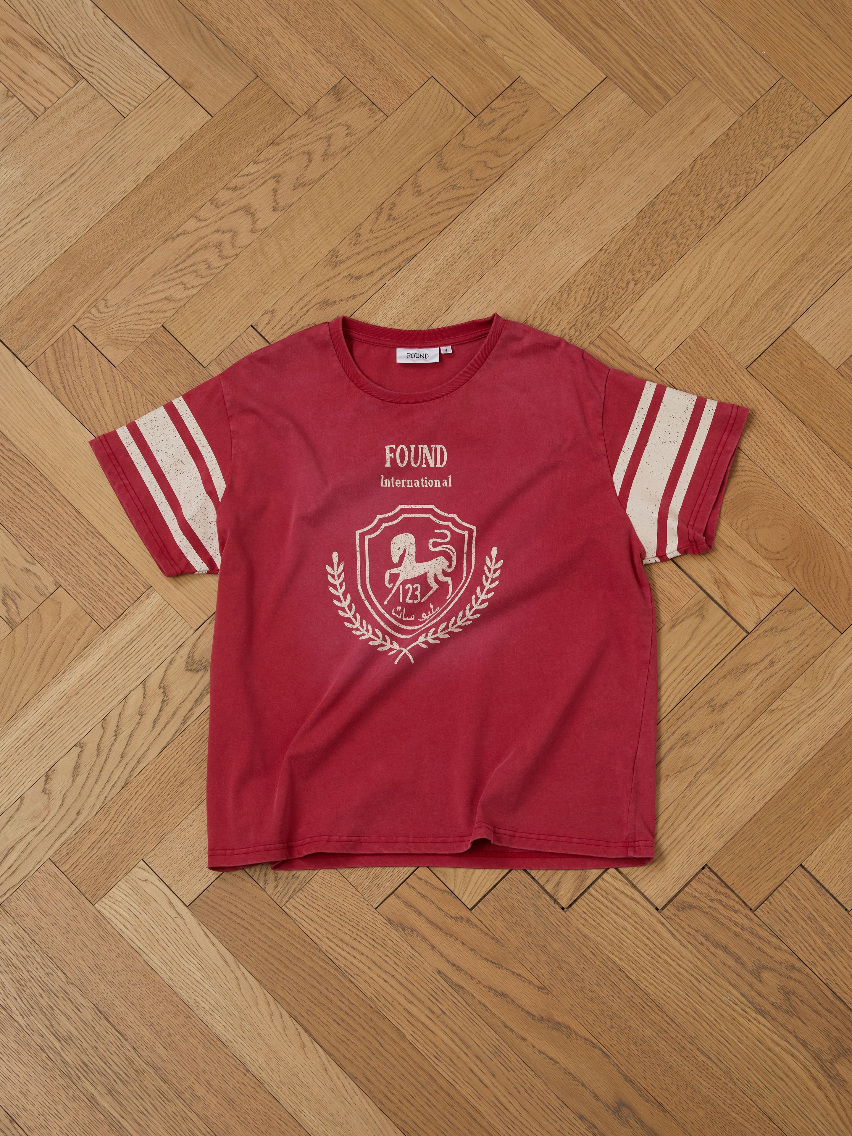 The Collegiate Crest Tee by FOUND is a unisex, relaxed fit red t-shirt with white sleeve stripes. Featuring "FOUND International" and a horse-emblazoned crest on the front, it exudes vintage athletic gear style, laid out on a wooden herringbone floor for display.