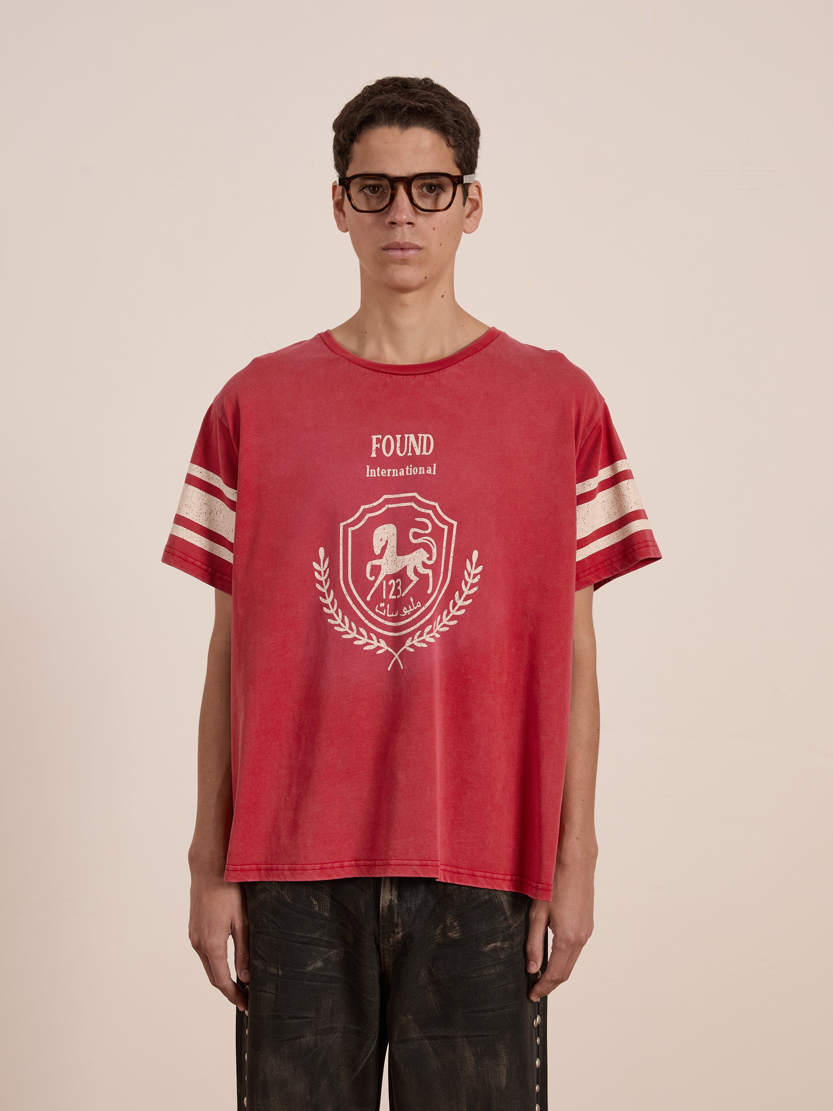 A person wearing glasses and a vintage FOUND Collegiate Crest Tee confidently stands in dark pants against a plain backdrop. The red tee's unisex relaxed fit, adorned with striped sleeves, subtly nods to classic athletic wear.