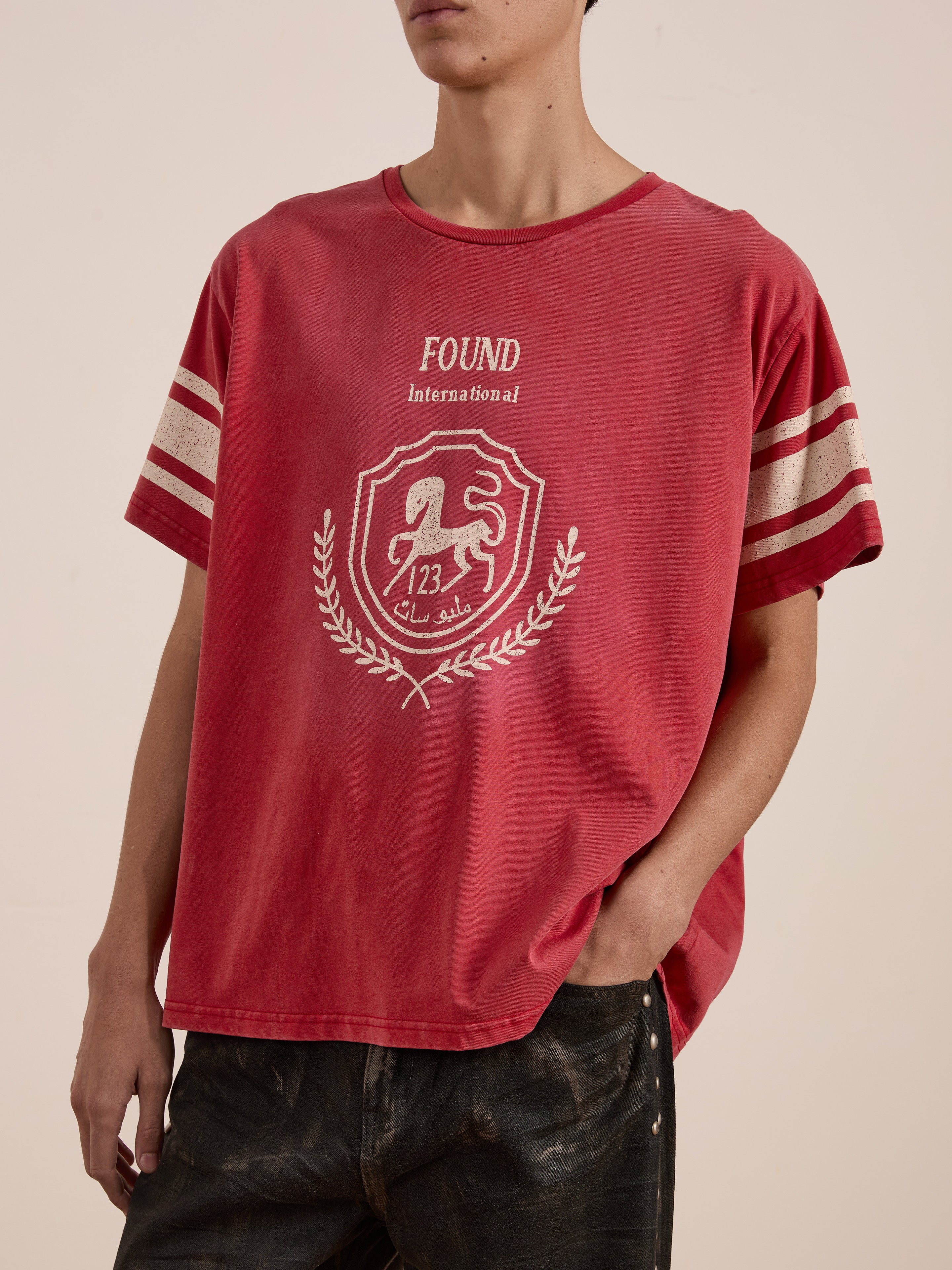 A person is wearing a red Collegiate Crest Tee by FOUND, with wide sleeves and featuring a crest logo and the words "FOUND International." This unisex relaxed fit garment nods to vintage athletic gear, combining classic style and modern comfort.