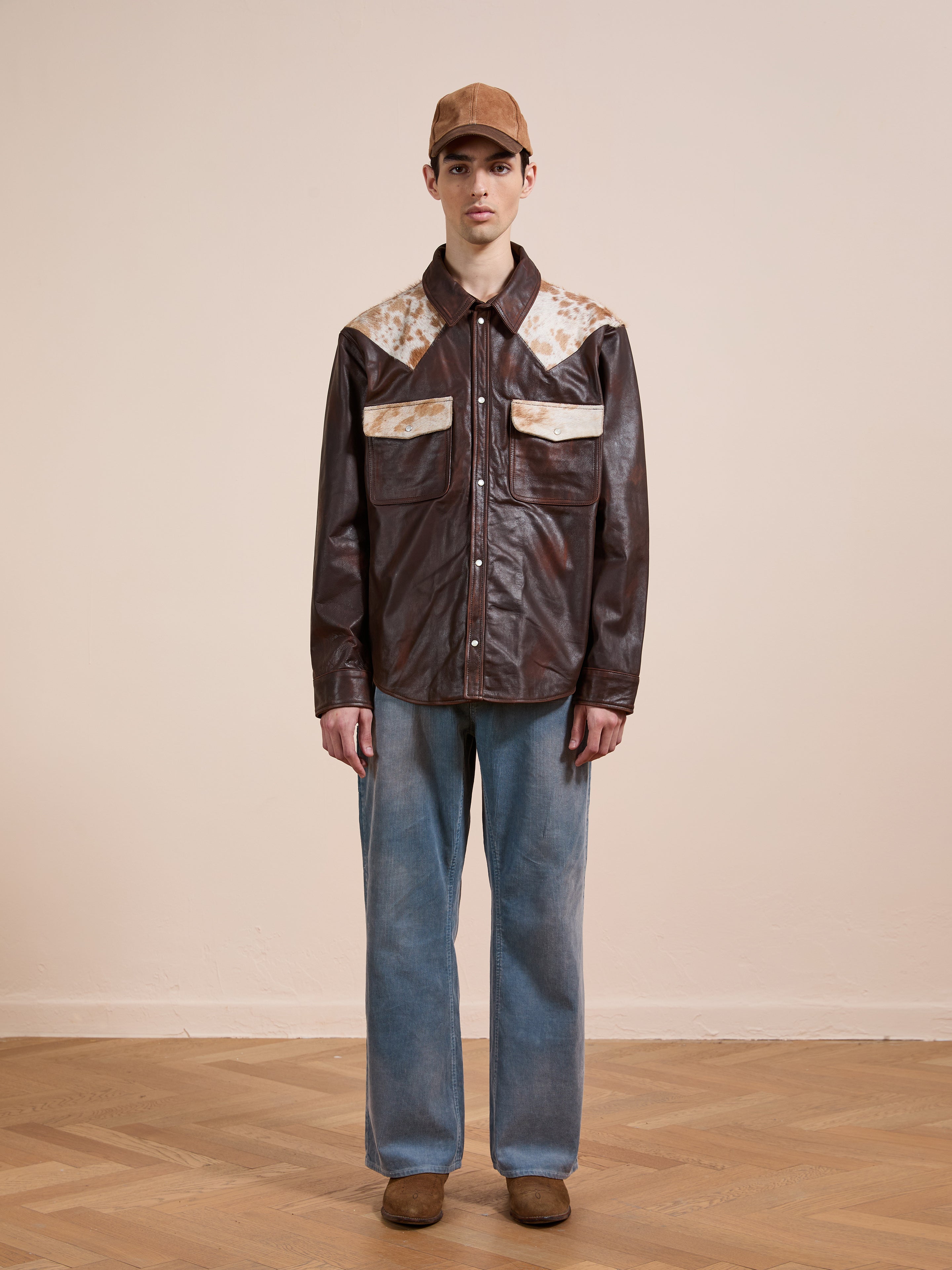 Indoors, a person wears the FOUND Distressed Leather Cowhide Contrast Western Shirt with blue jeans, brown shoes, and a brown cap. The genuine cowhide shirt offers a unisex relaxed fit for easy, casual style.