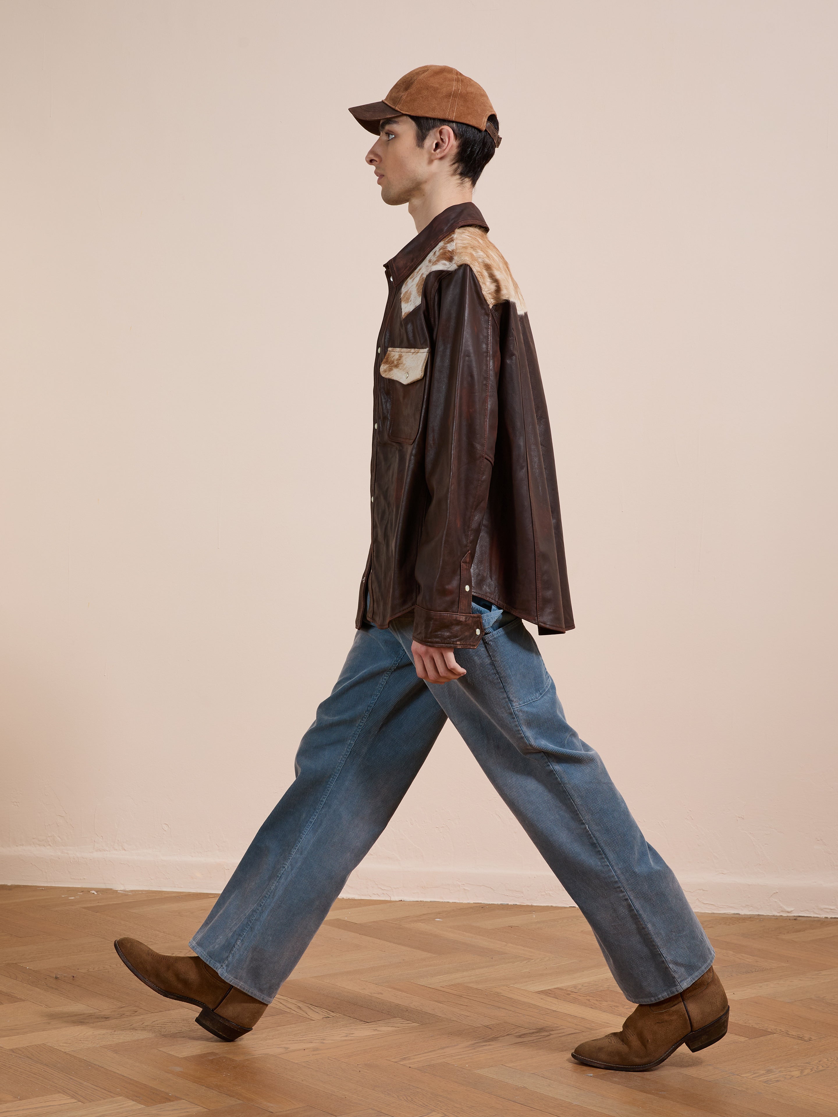 A man in a brown cap, FOUND Distressed Leather Cowhide Contrast Western Shirt, blue jeans, and brown boots walks indoors on a wooden floor against a plain beige wall.