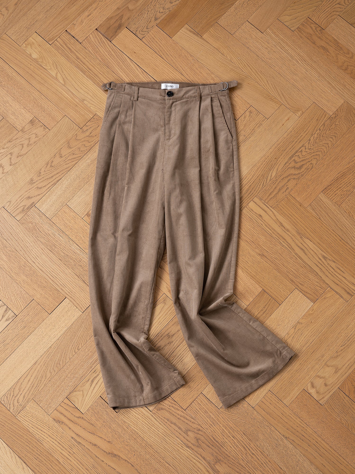 A pair of FOUND's Corduroy Pleated Trousers in brown, featuring a high-rise waist, lies on a wooden herringbone-patterned floor.